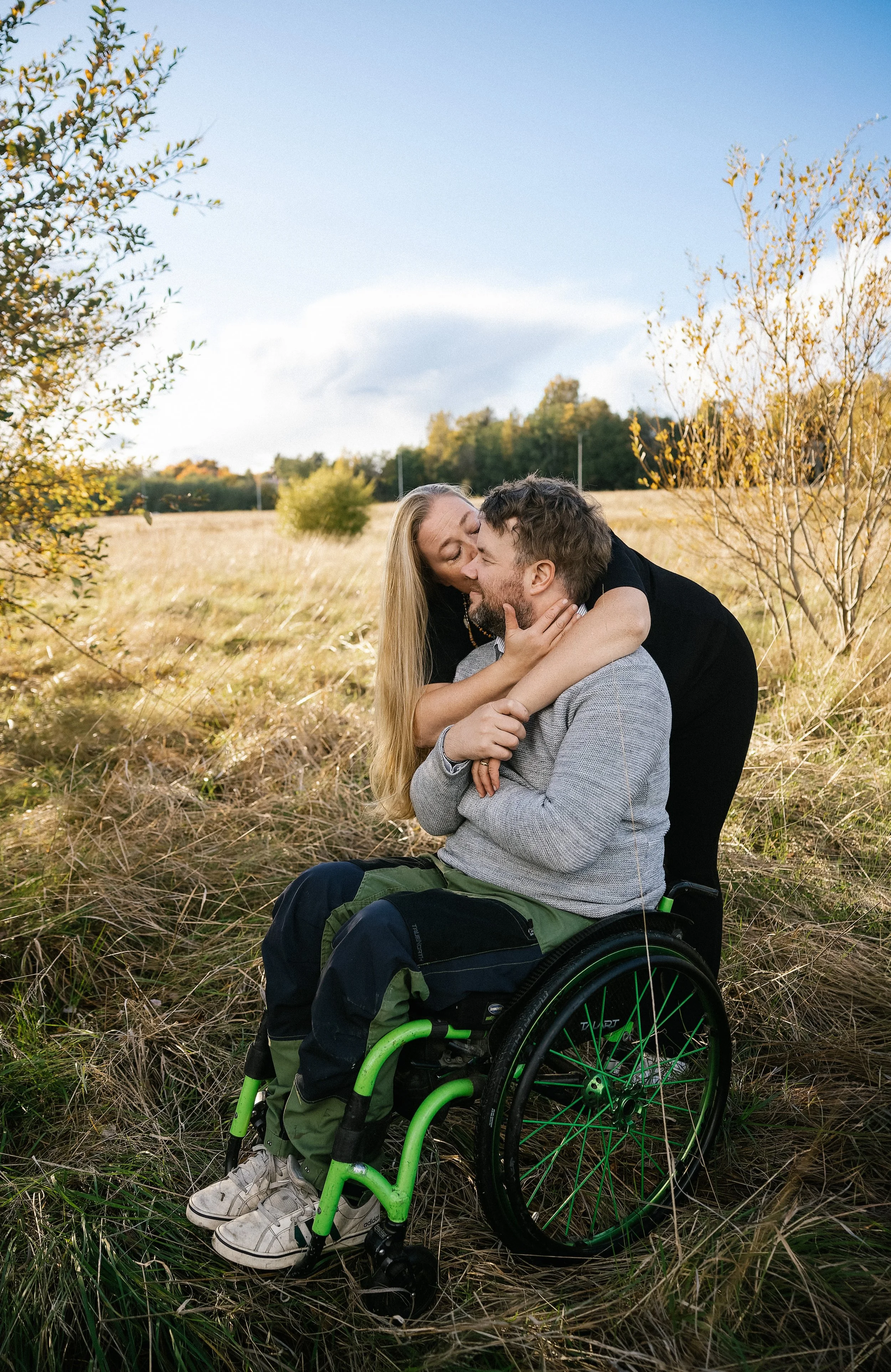 A woman and a man in a wheelchair sharing a kiss outdoors in a grassy field with trees and blue sky in the background during fall.