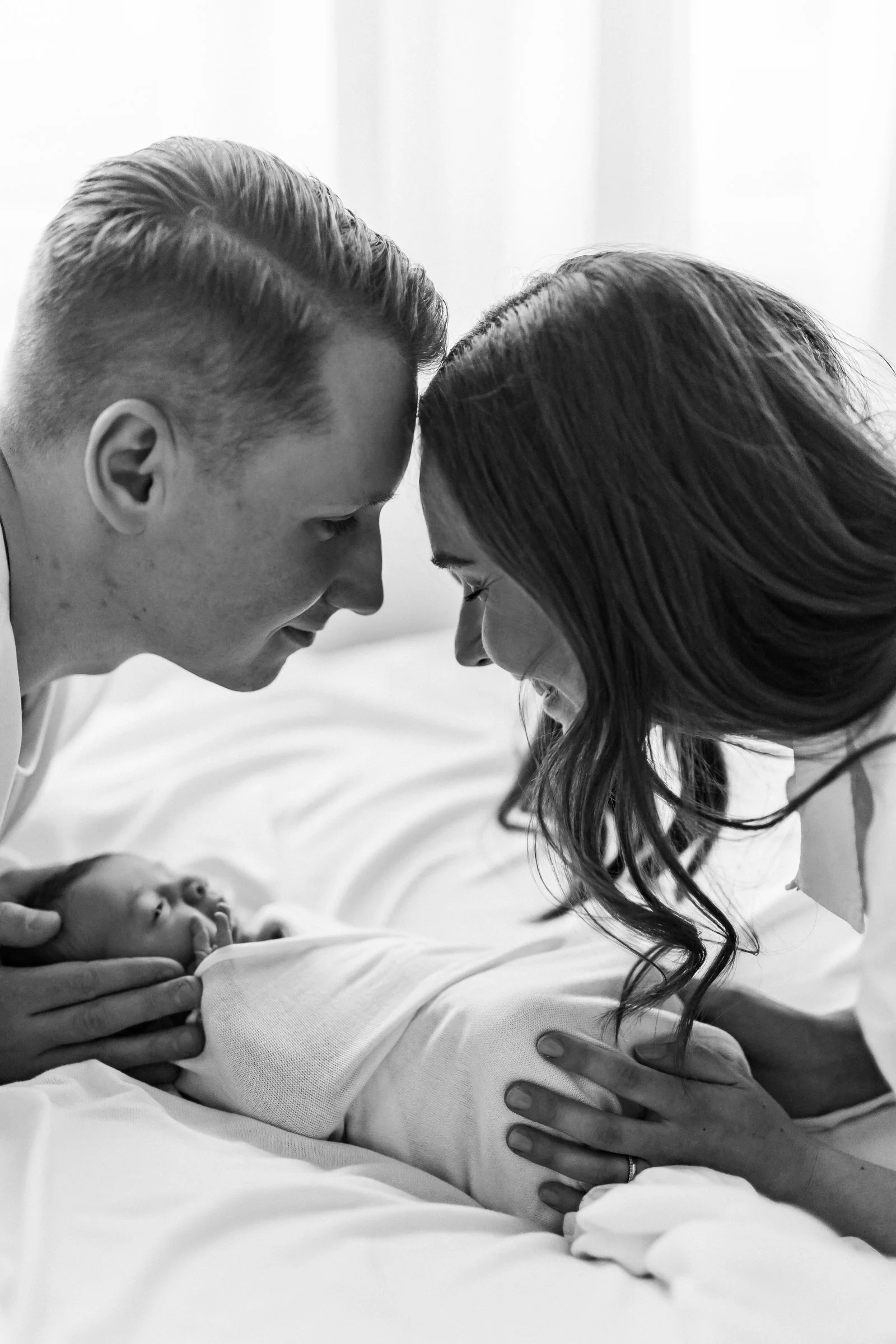 A black and white photo of a new family with a newborn baby. The father and mother are touching foreheads and smiling, while the mother is holding the baby's head gently, feeling the baby's tiny hand.
