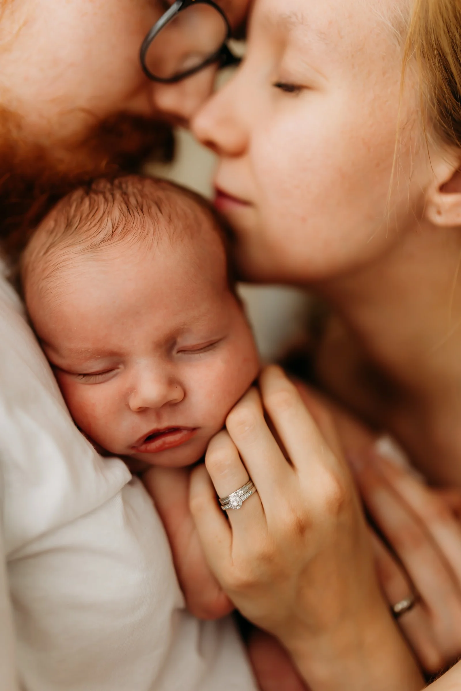 Close-up of a woman, a man, and a baby, with the woman and man gently touching the baby's face and the man leaning in to kiss the woman on the forehead, all with peaceful expressions.