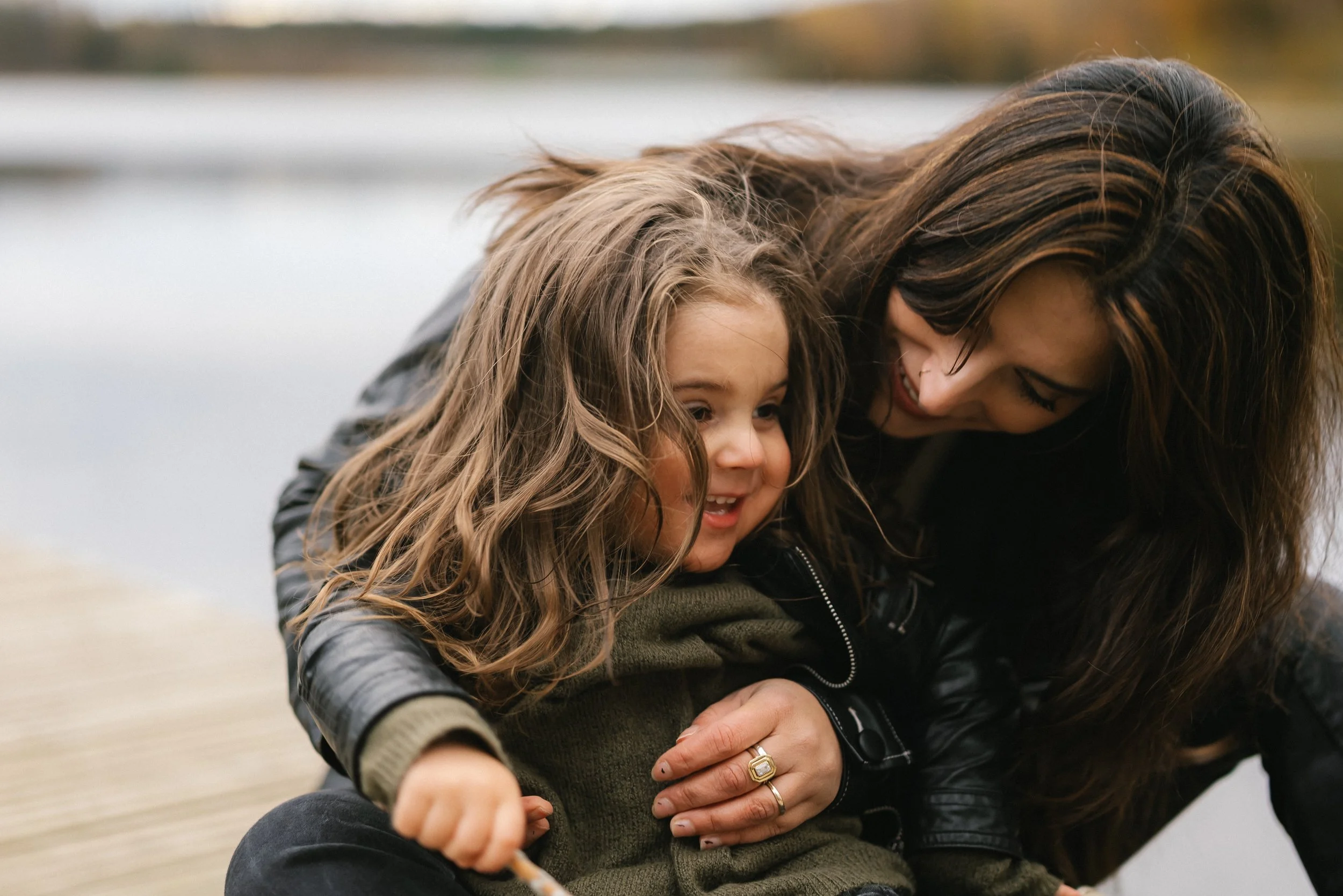 A woman and a young girl playing together outdoors on a wooden dock by a body of water, smiling and laughing.