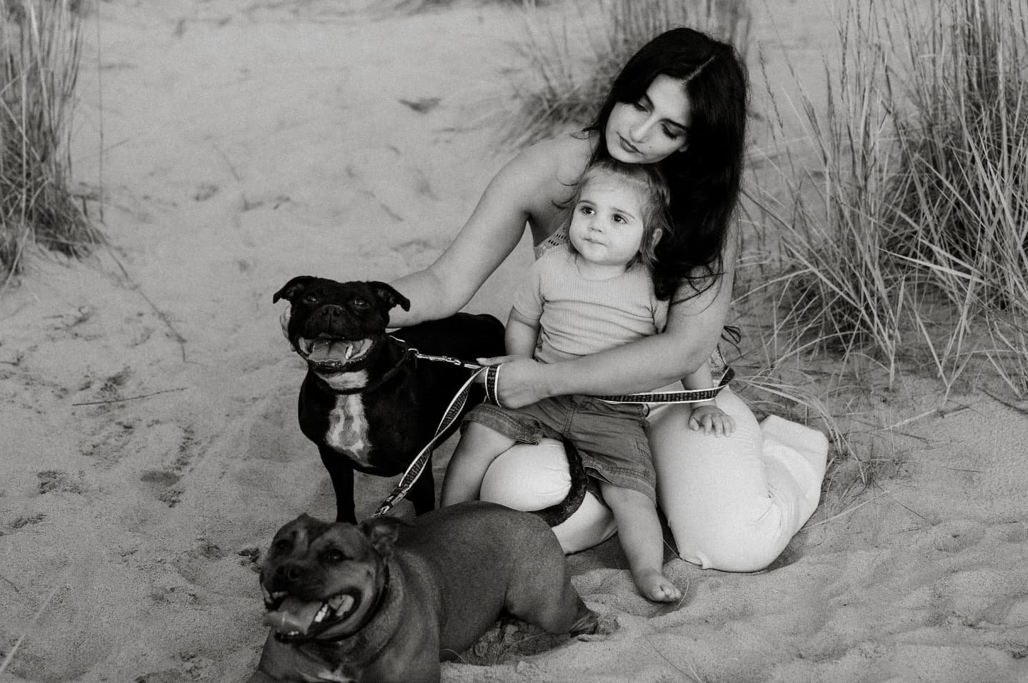A woman and a young girl sitting on the sand at the beach, surrounded by two dogs, with grassy dunes in the background.