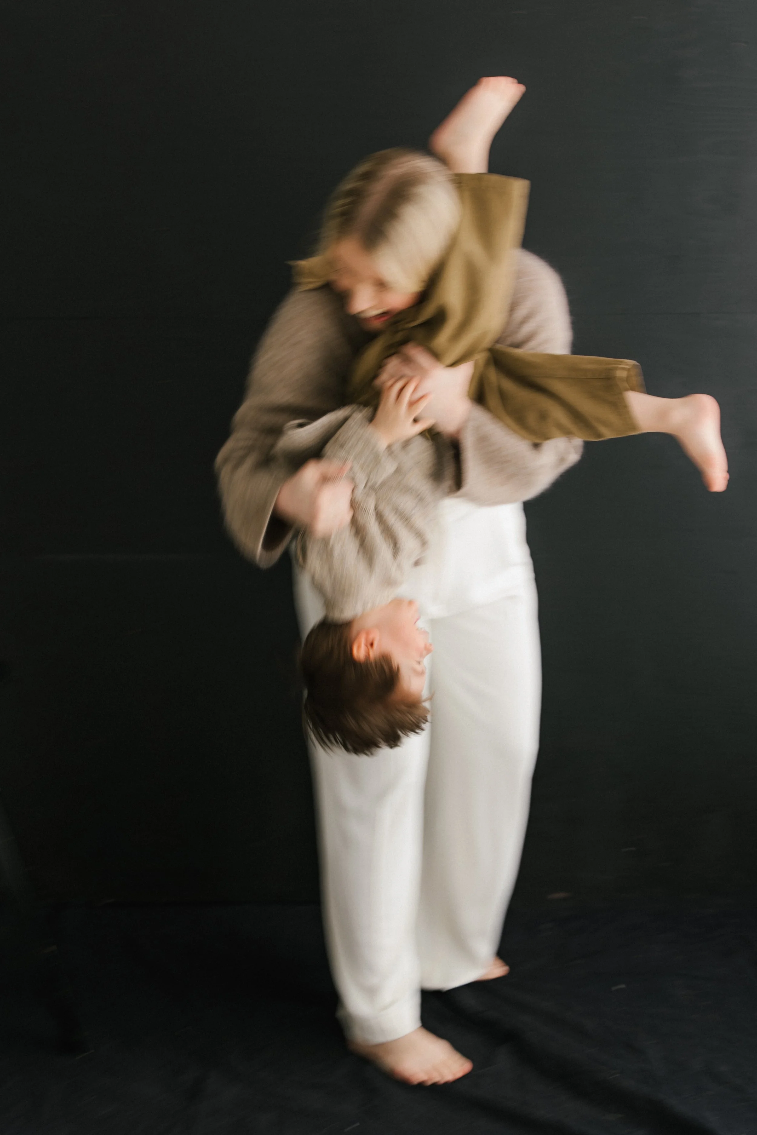 A woman with shoulder-length blonde hair is playing with two children against a black background. One child is upside down, and the other is being held and appears to be smiling.