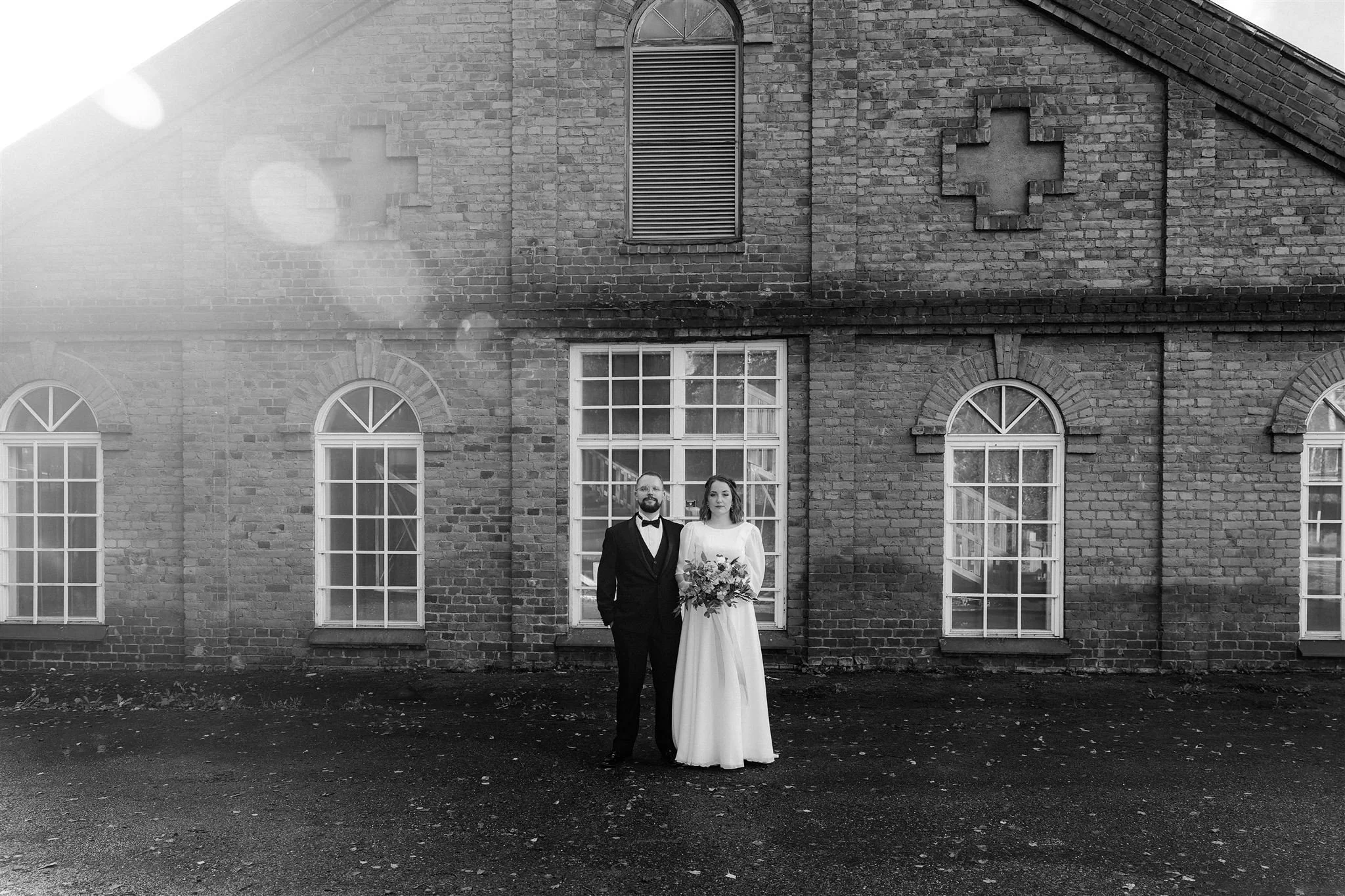 A couple in wedding attire standing in front of a brick building with arched windows, one holding a bouquet of flowers, on a sunny day.