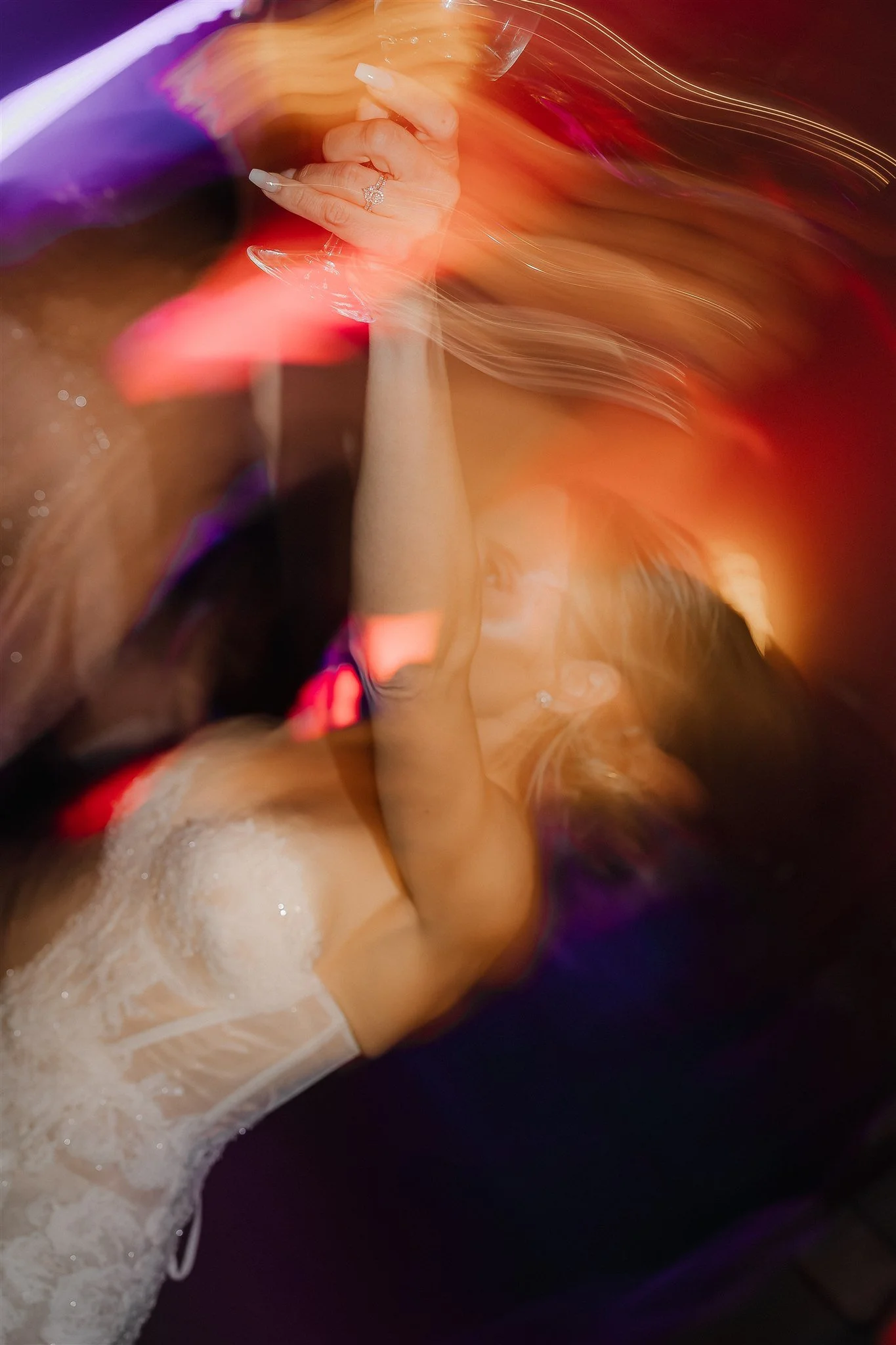 A woman dancing in a colorful, blurred light-filled setting, wearing a white lace dress and holding a glass of wine.