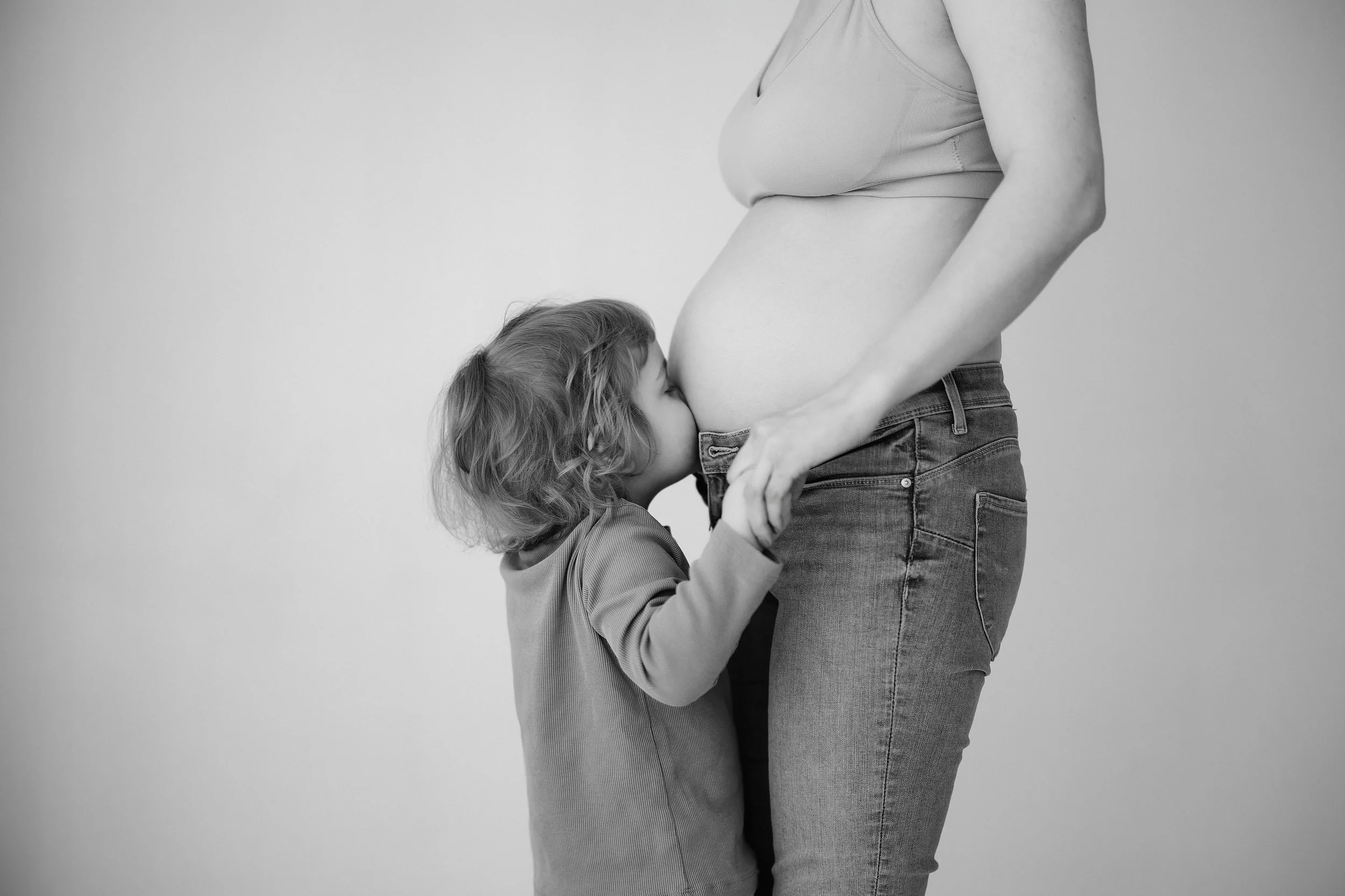 A young girl kisses a pregnant woman's belly, which is visible as she wears jeans and a sleeveless top, against a plain background.