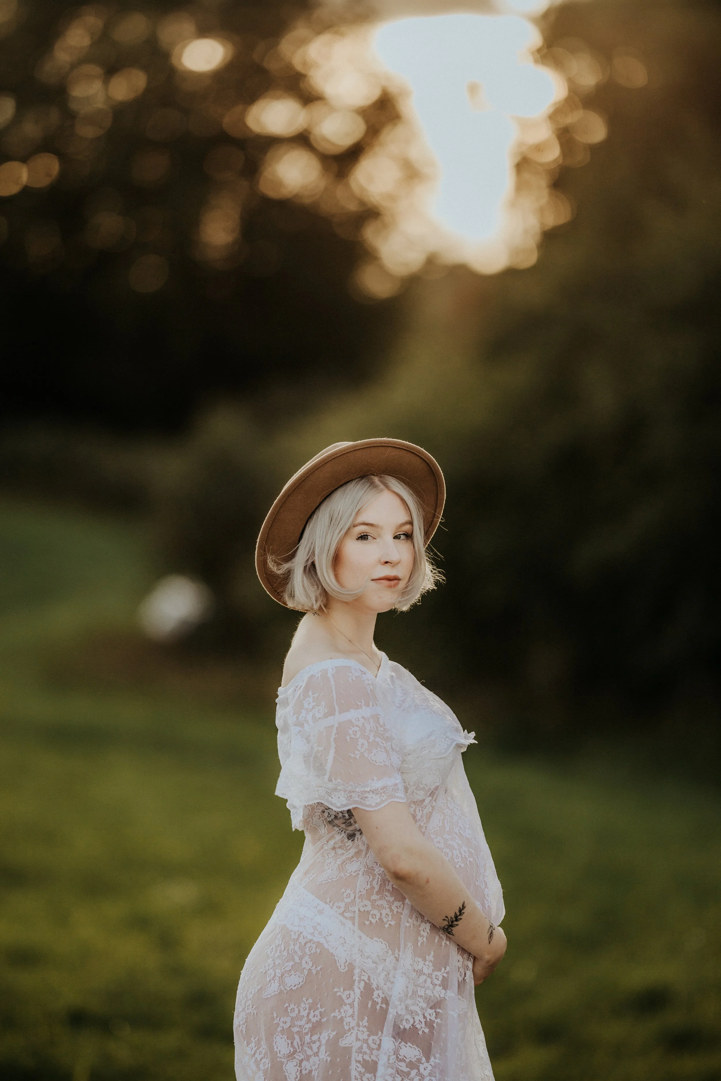 A young woman with blonde hair wearing a brown hat and a sheer white lace dress standing outdoors during sunset.