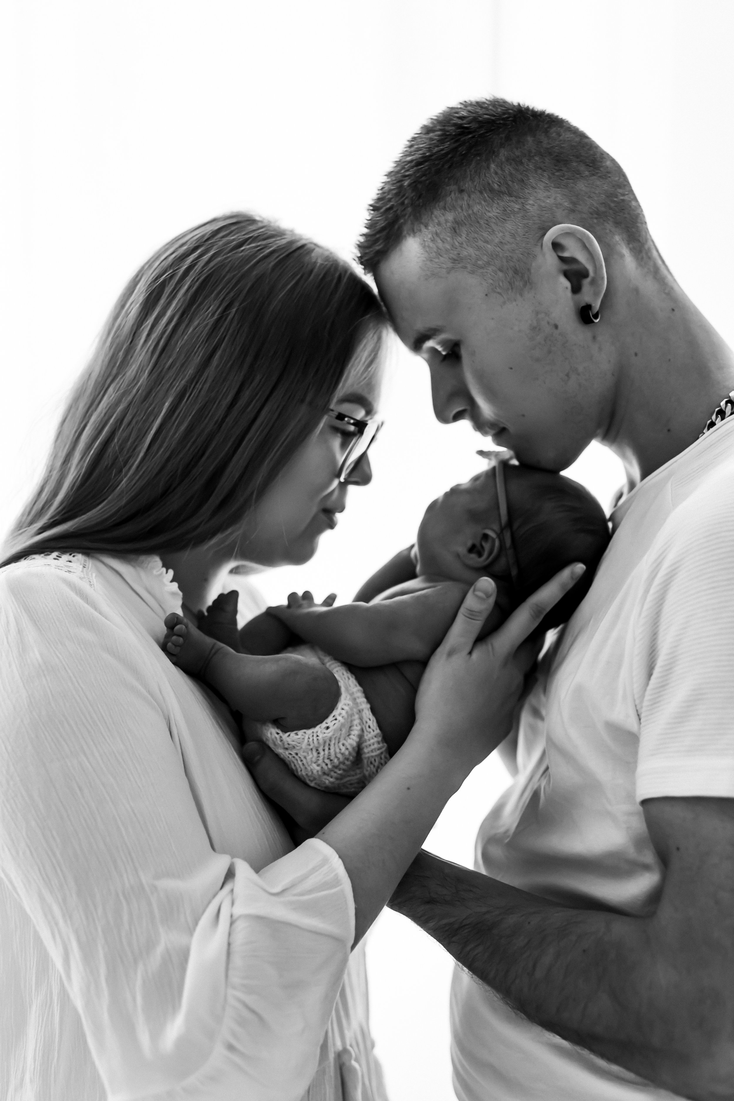 A black and white photo of a couple holding their newborn baby close to their faces, with the mother wearing glasses and the father with short hair and earrings.