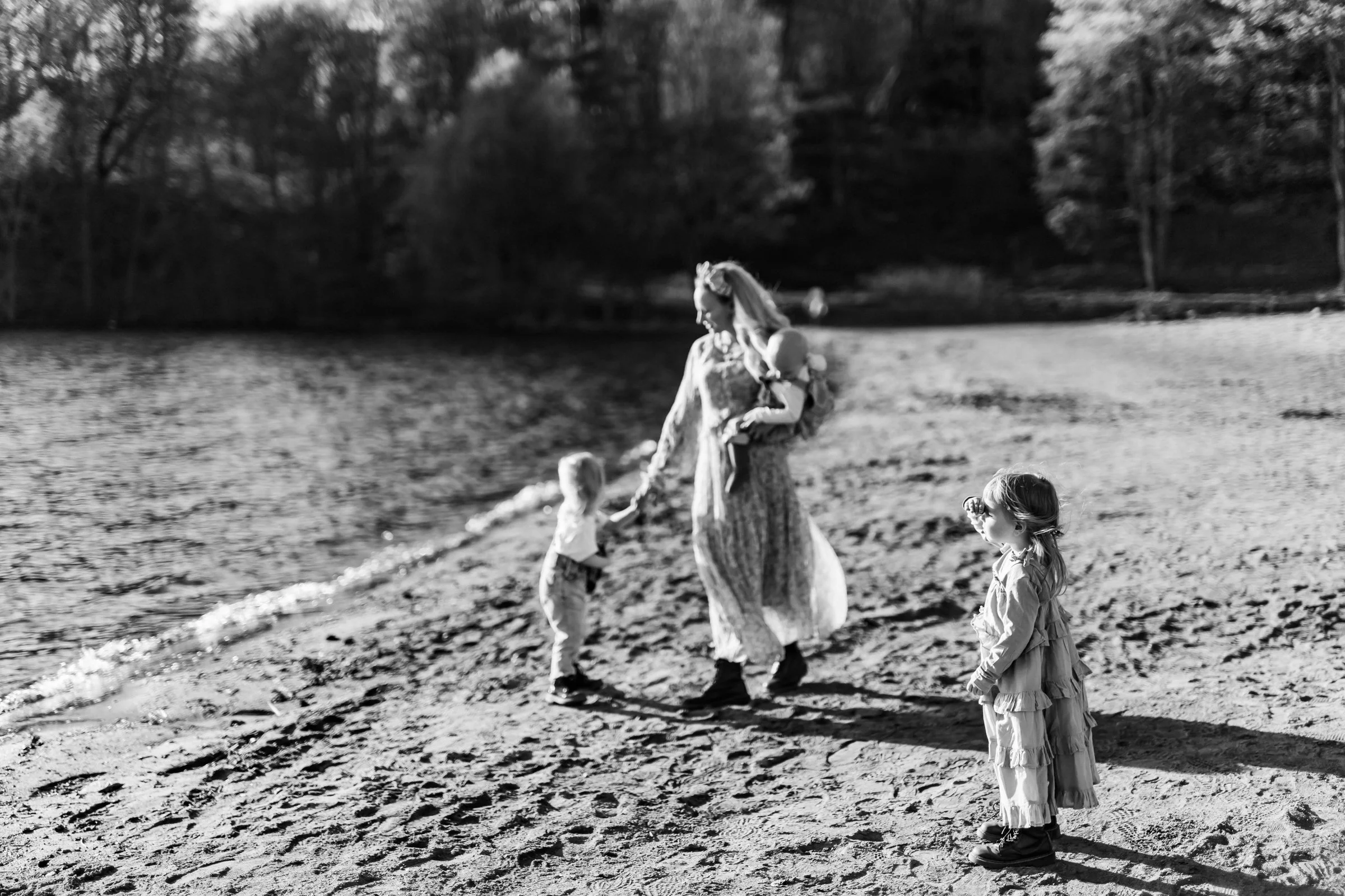 A woman holding a young girl and a young boy holding her hand on a beach near the water, with trees in the background.