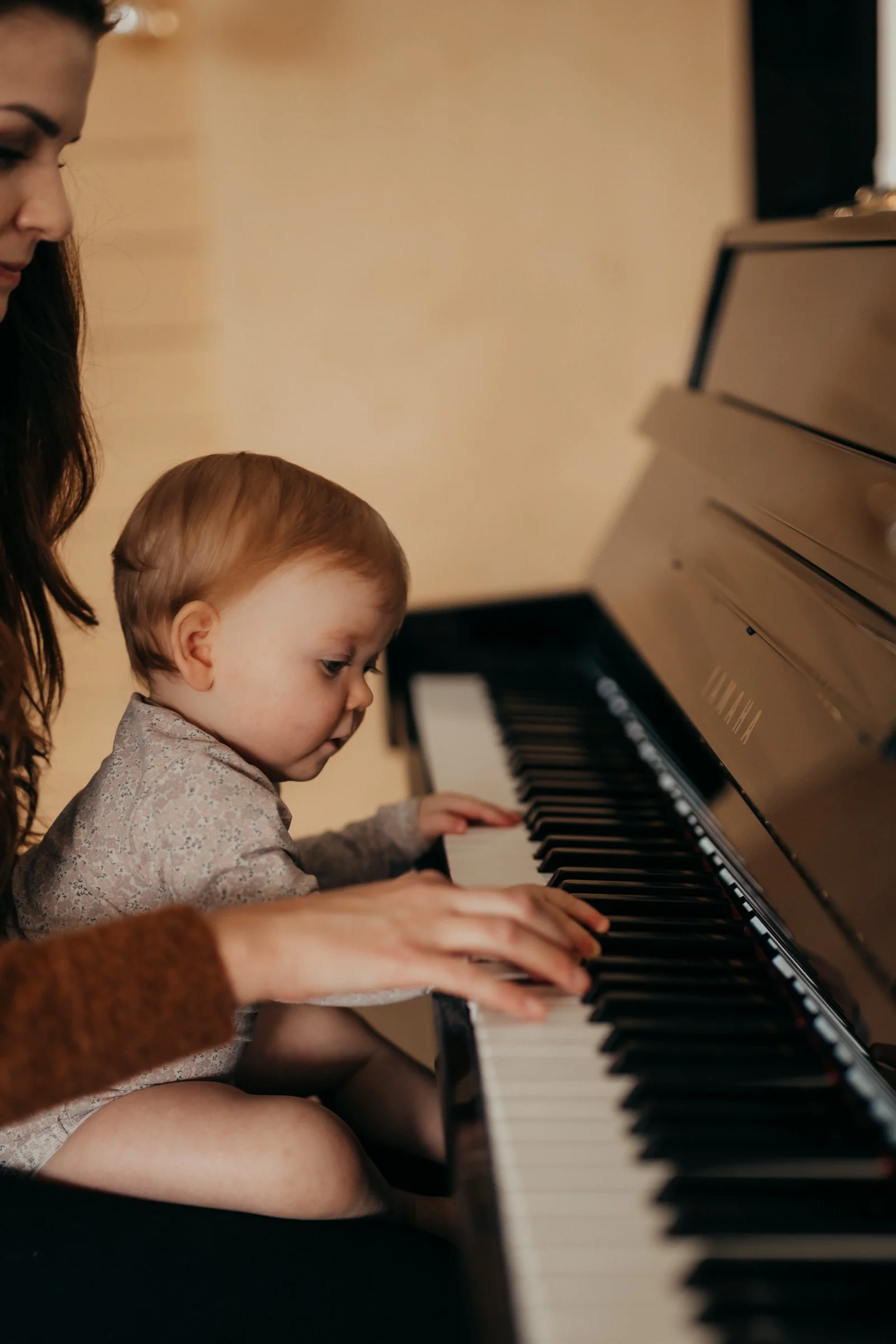 A young girl and a toddler sitting at a piano, with the girl guiding the child's hands on the keys.