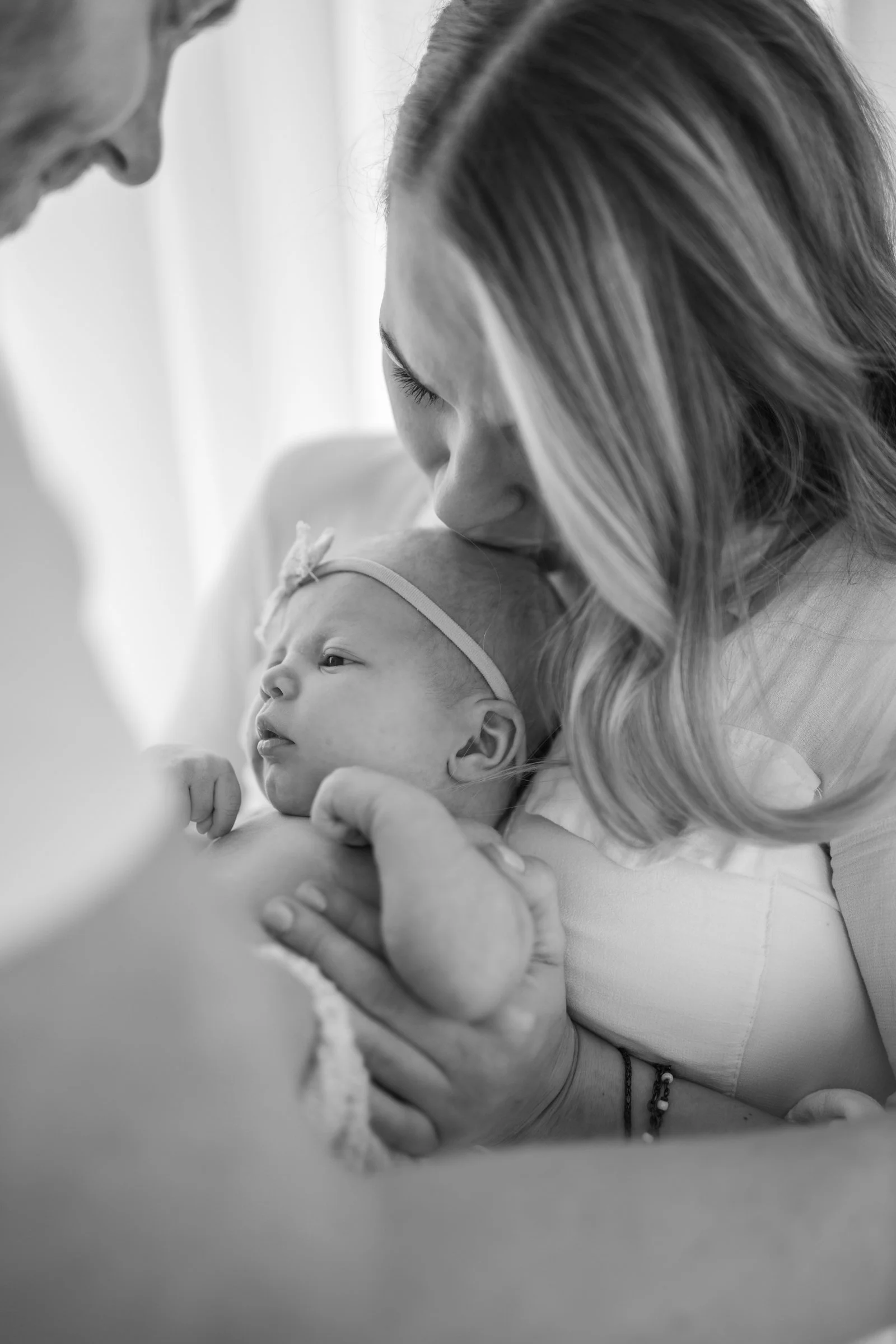 Black and white photo of a woman kissing a newborn baby on the head, held in her arms, with a man looking on.