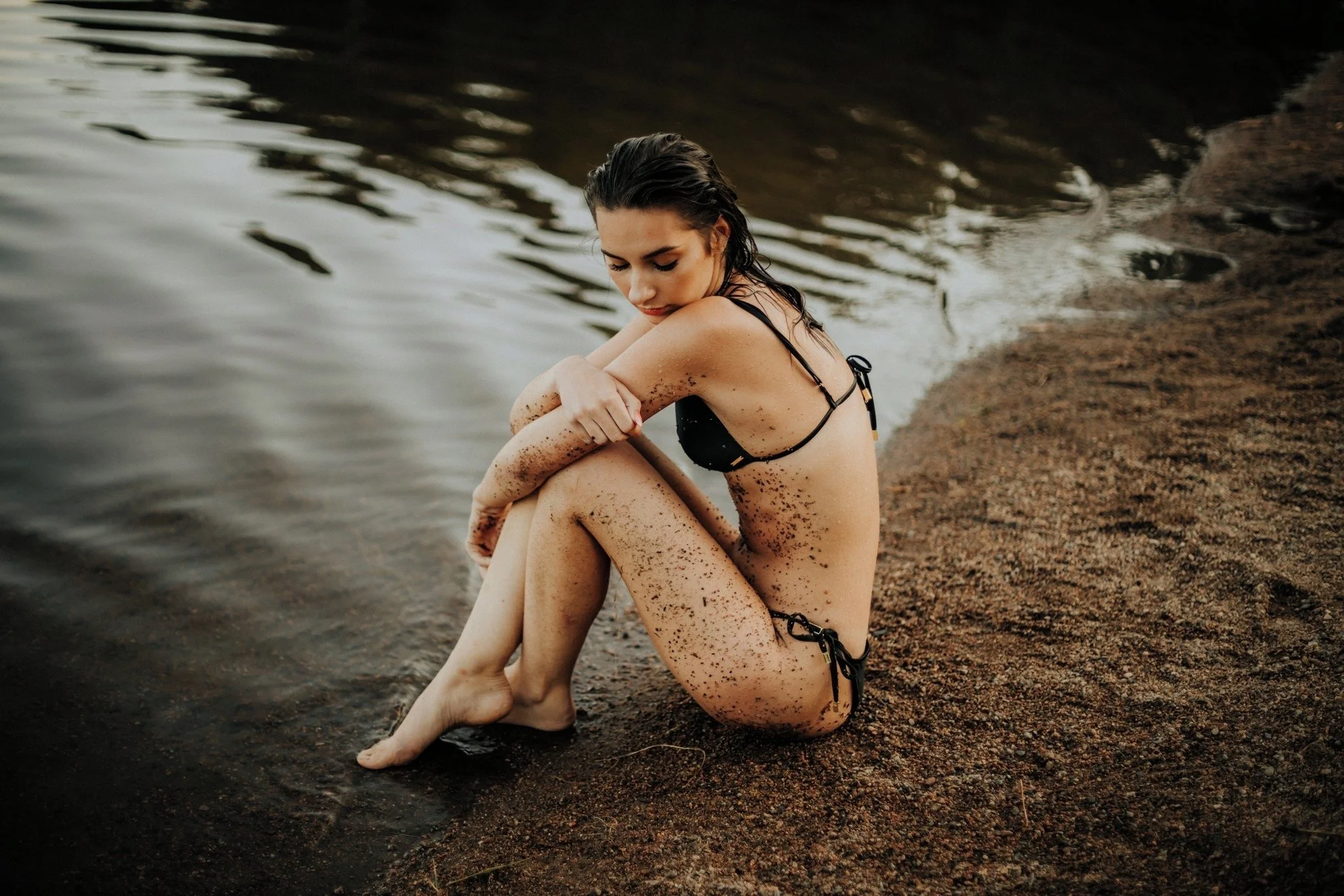 A woman sitting on the sandy shore of a body of water, with her arms wrapped around her knees and her head bowed, wearing a black bikini, with wet hair and sand on her skin.