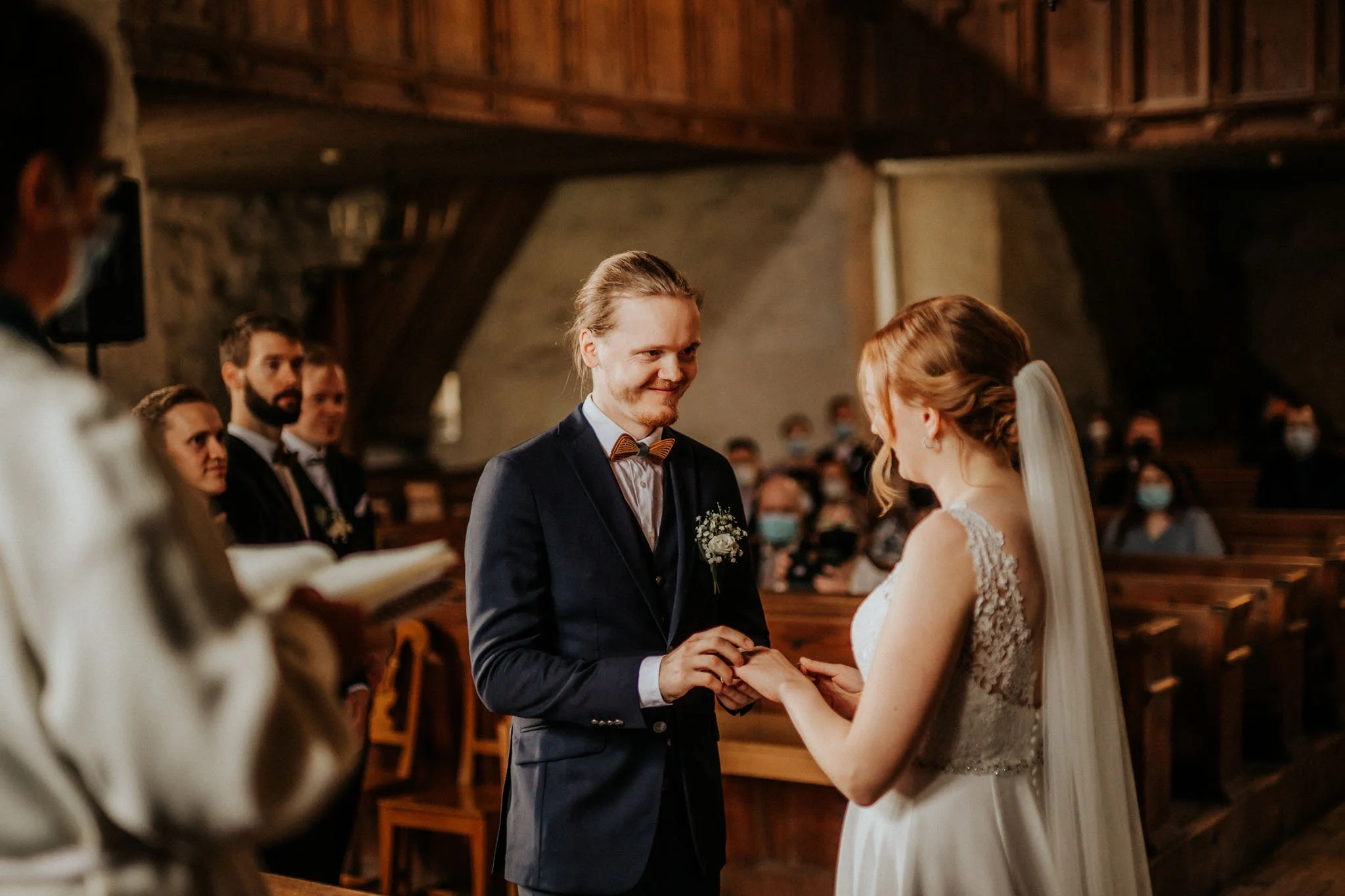 A couple getting married at a church ceremony, with the bride in a white lace dress and the groom in a dark suit, exchange rings while guests watch.