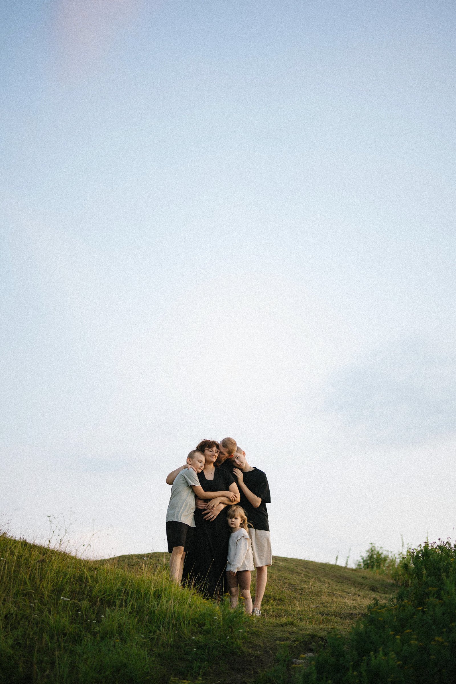 A group of five people, including children and an adult woman, standing closely together outdoors on a grassy hill at sunset, embracing and looking content.