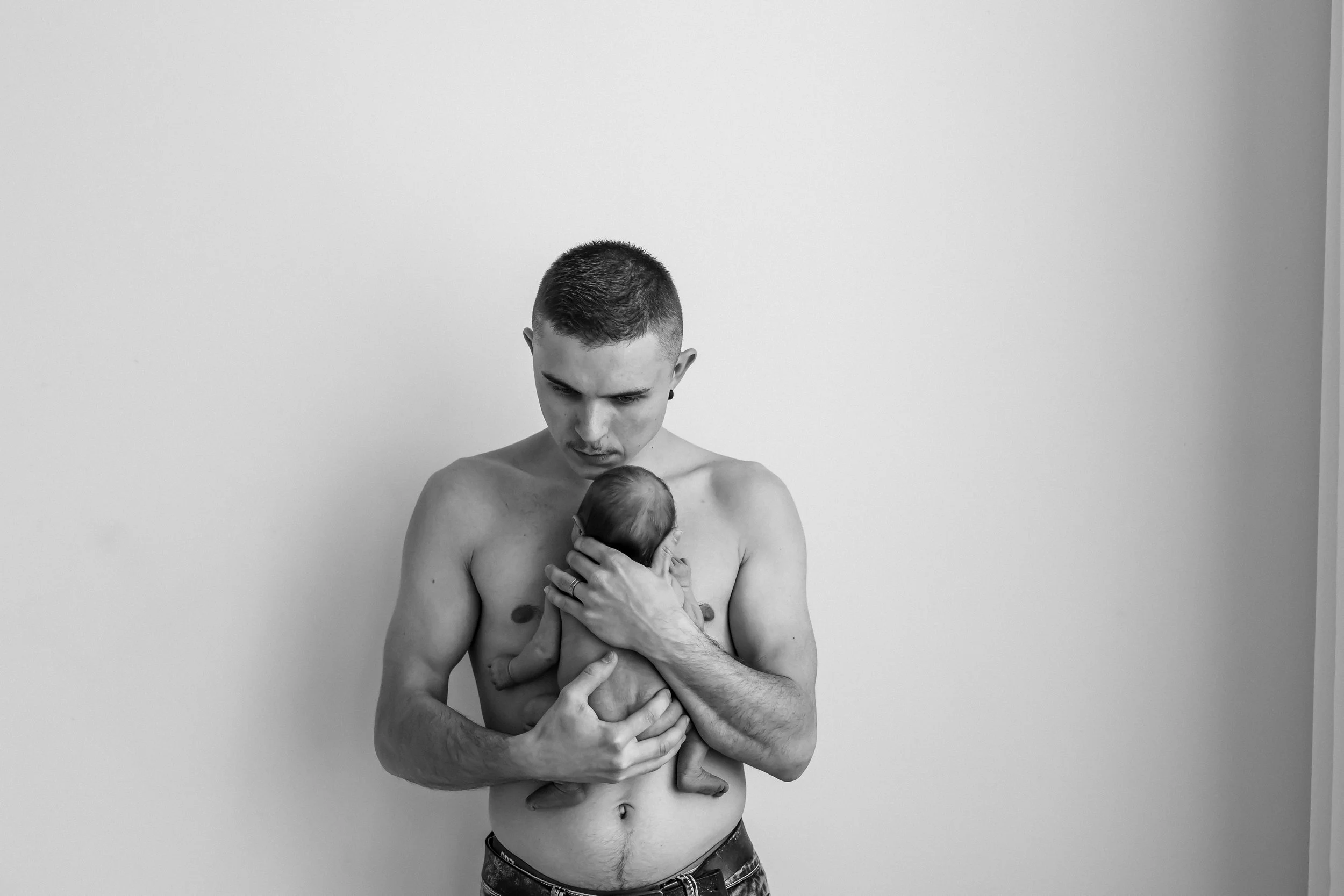 Black and white photo of a shirtless man holding a small baby against a plain wall.