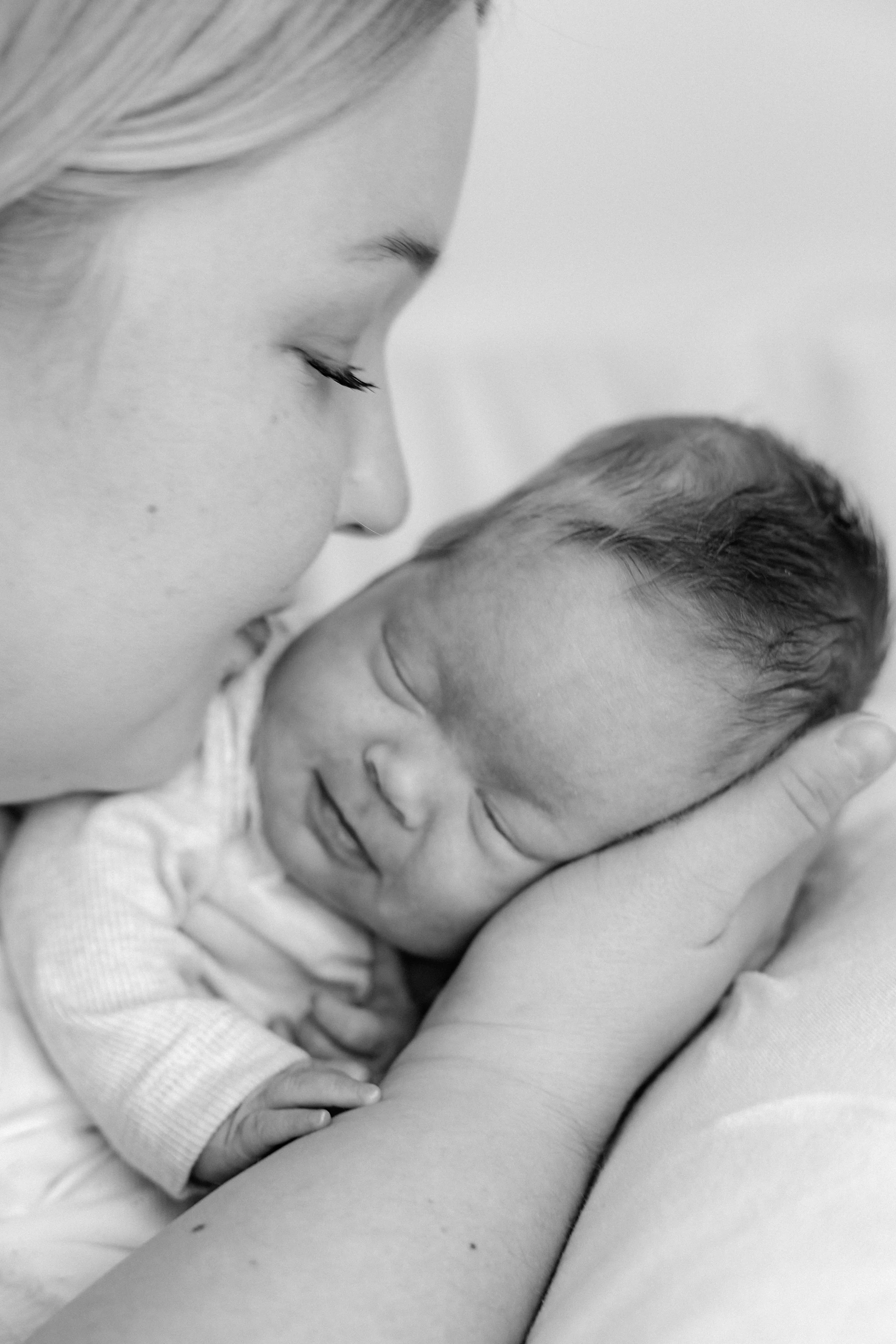 A woman is holding a sleeping newborn baby close to her face, with her eyes closed, creating an intimate and tender moment.