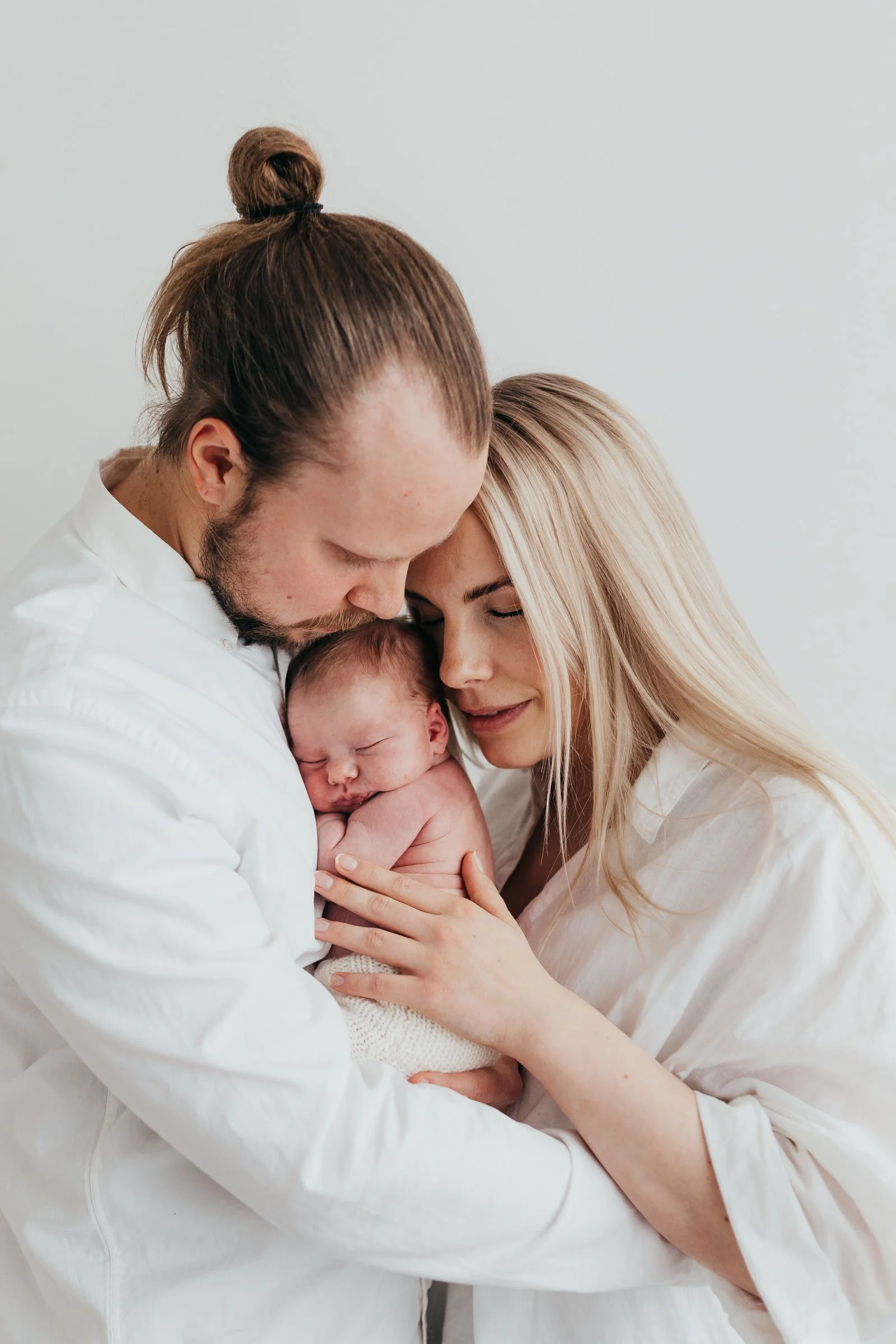 A newborn baby held by a man with a top knot hairstyle and a woman with long blonde hair, all with closed eyes in a tender moment.