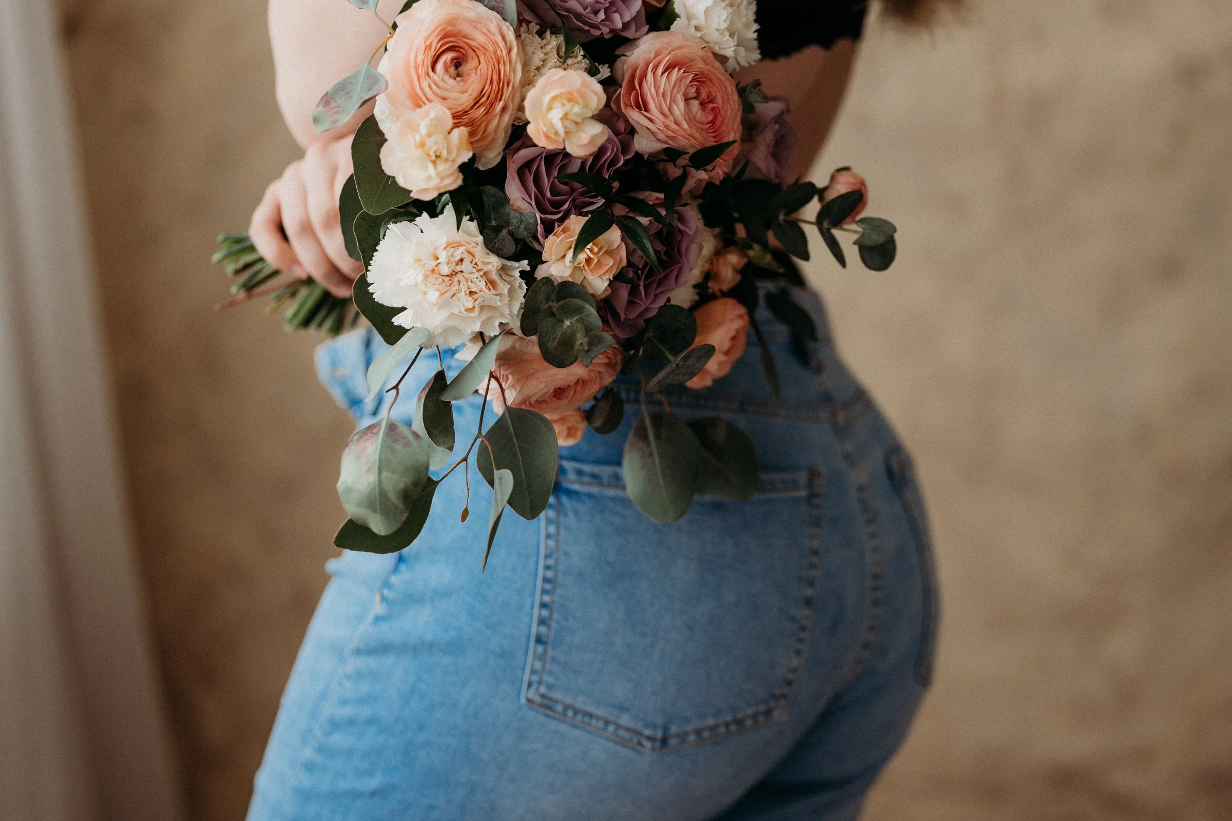 Person holding a bouquet of peach, white, and purple flowers with green leaves, wearing blue jeans.