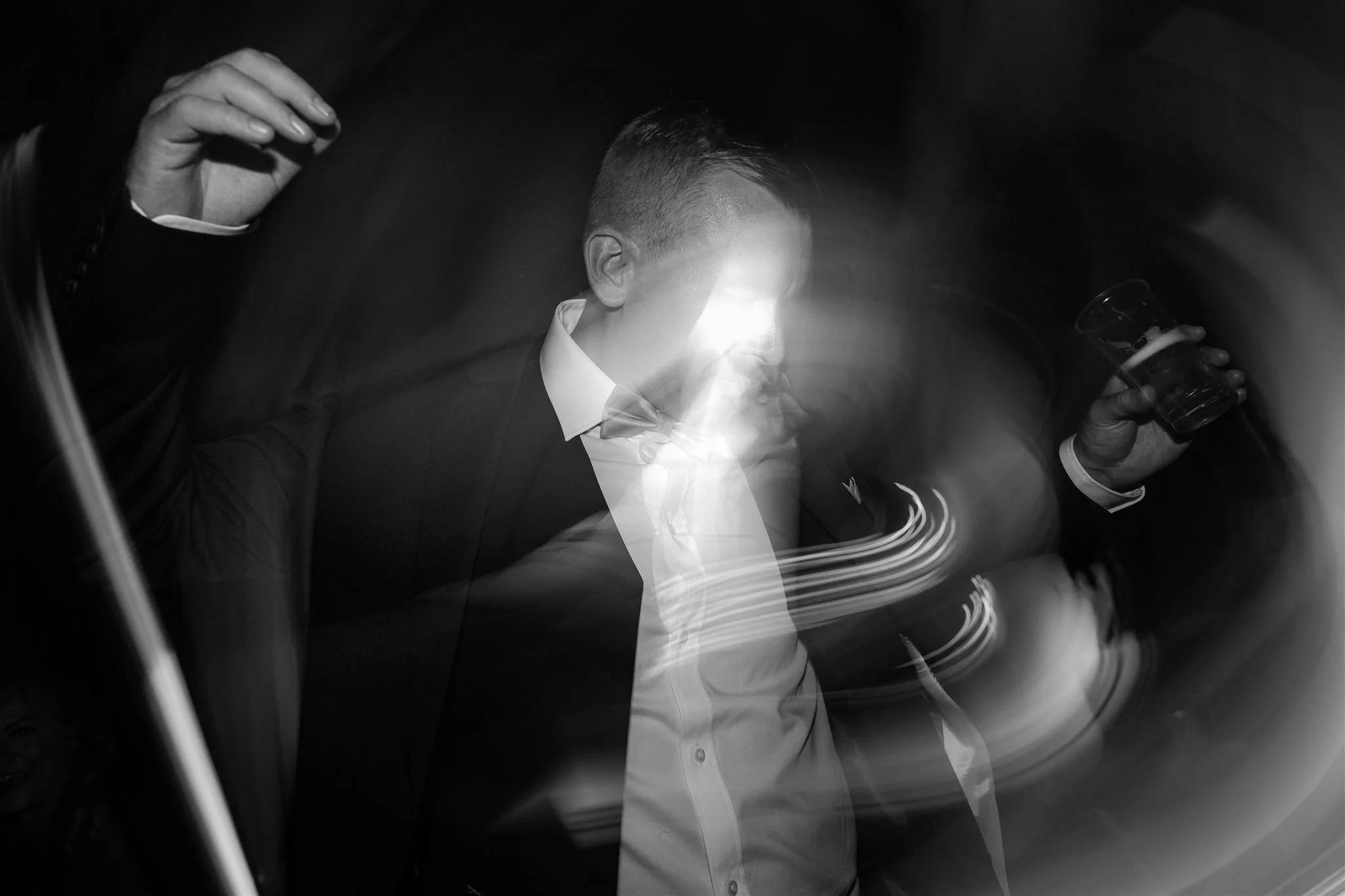 A black and white photo of a man in a suit holding a glass, with blurred motion and light streaks around him, emphasizing movement and a dynamic atmosphere.