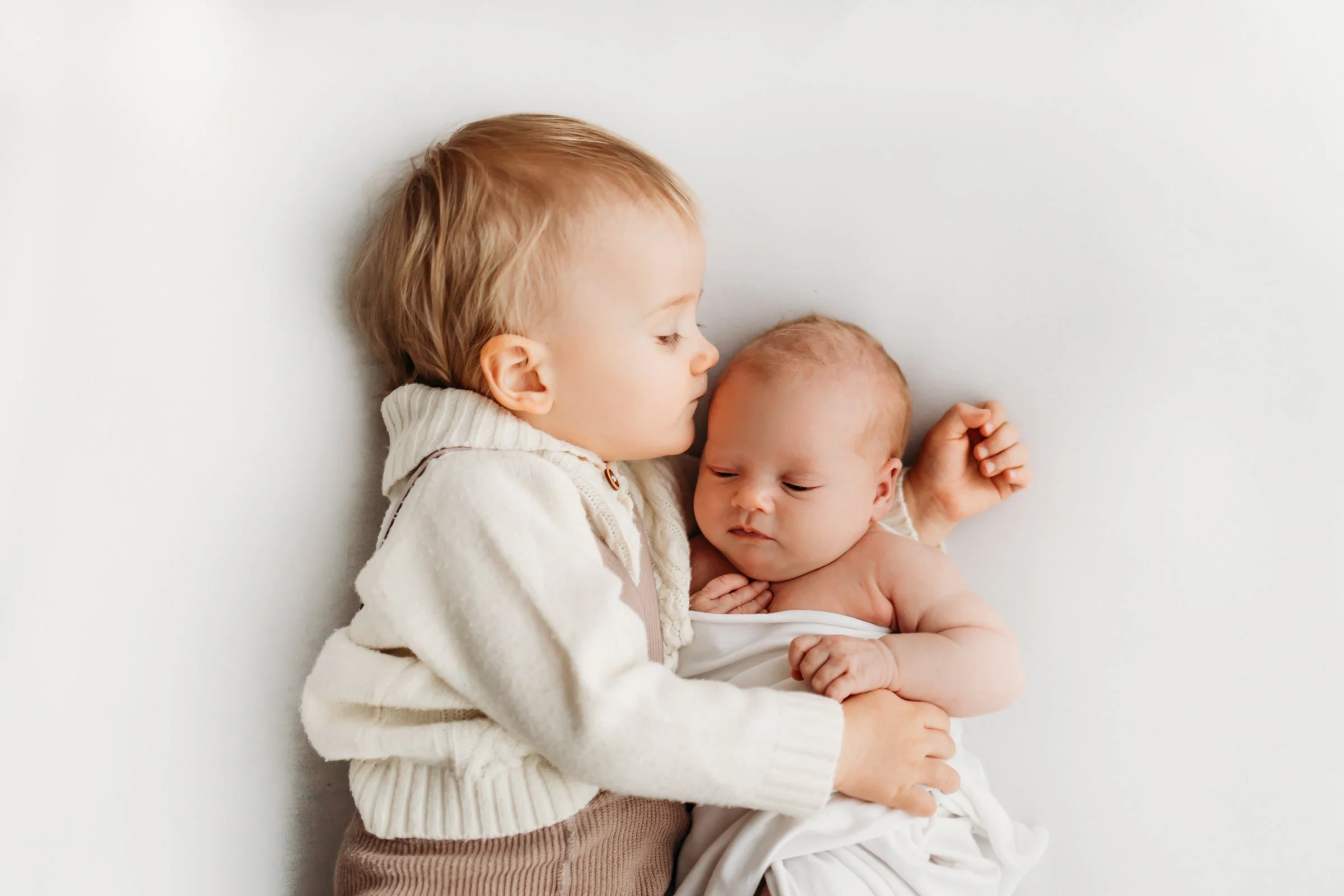 A young child and a newborn baby lying on a white surface, the older child gently sleeping while cradling the baby.