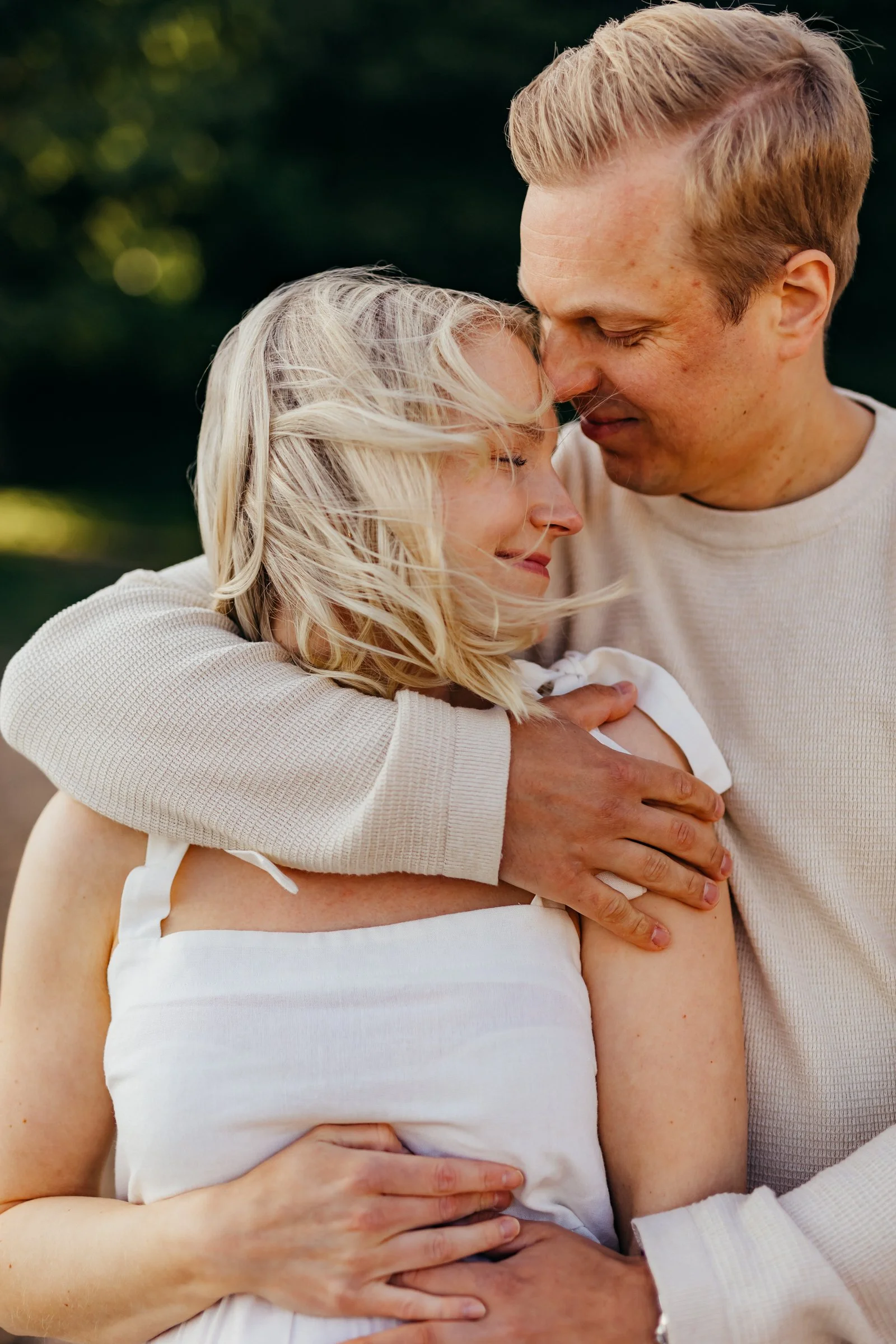 A couple embracing outdoors, forehead to forehead, with closed eyes and gentle smiles, in a close, intimate pose.