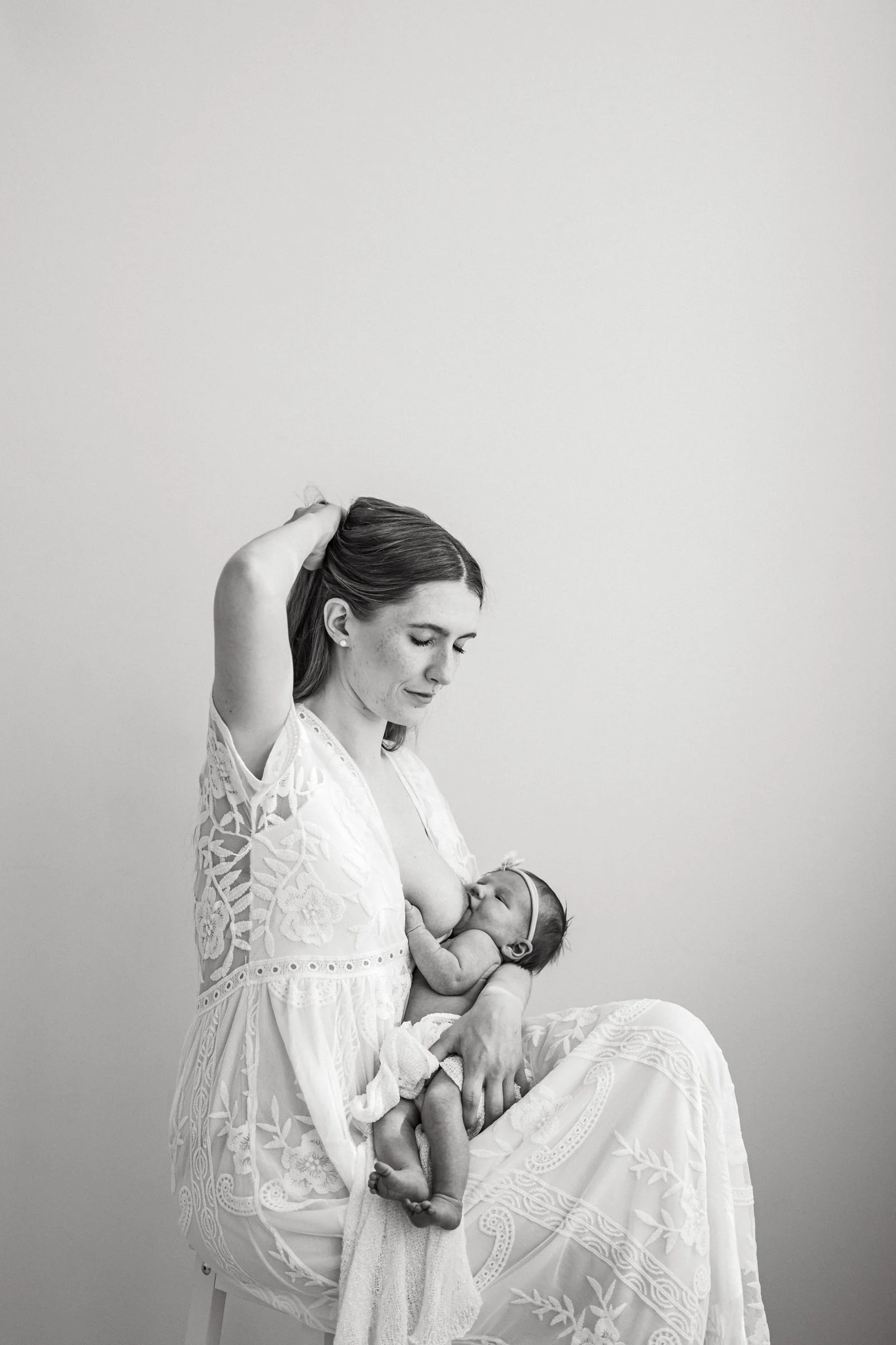 A woman is breastfeeding a baby while seated. The woman has her hand in her hair and is looking down lovingly at the baby. The baby is comfortably positioned in her lap, with a small bandage on its head, and is nursing.