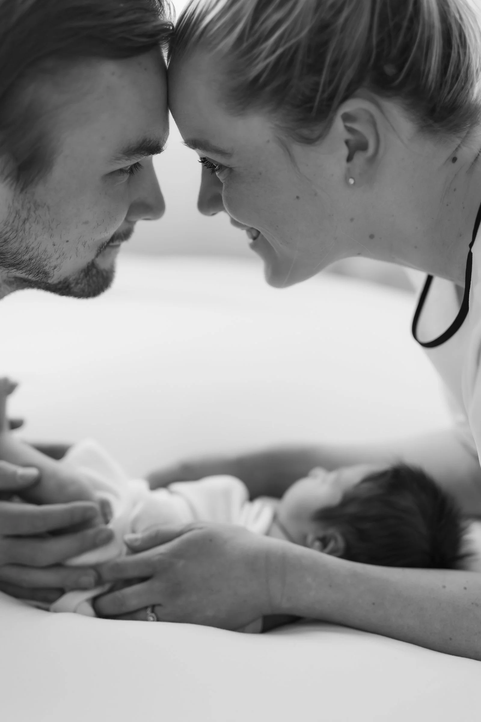 Black and white photo of a couple with foreheads touching, looking at each other, with a newborn baby lying on a bed beneath them.