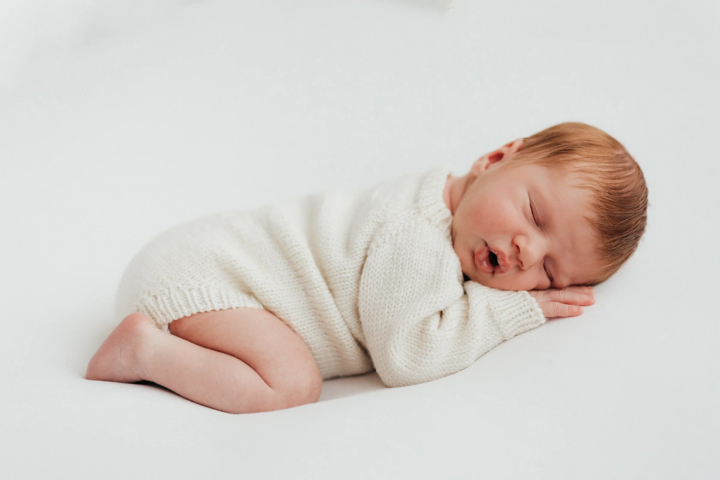 A sleeping baby with reddish-brown hair, lying on a white surface, wearing a cream-colored knitted sweater and matching diaper cover.