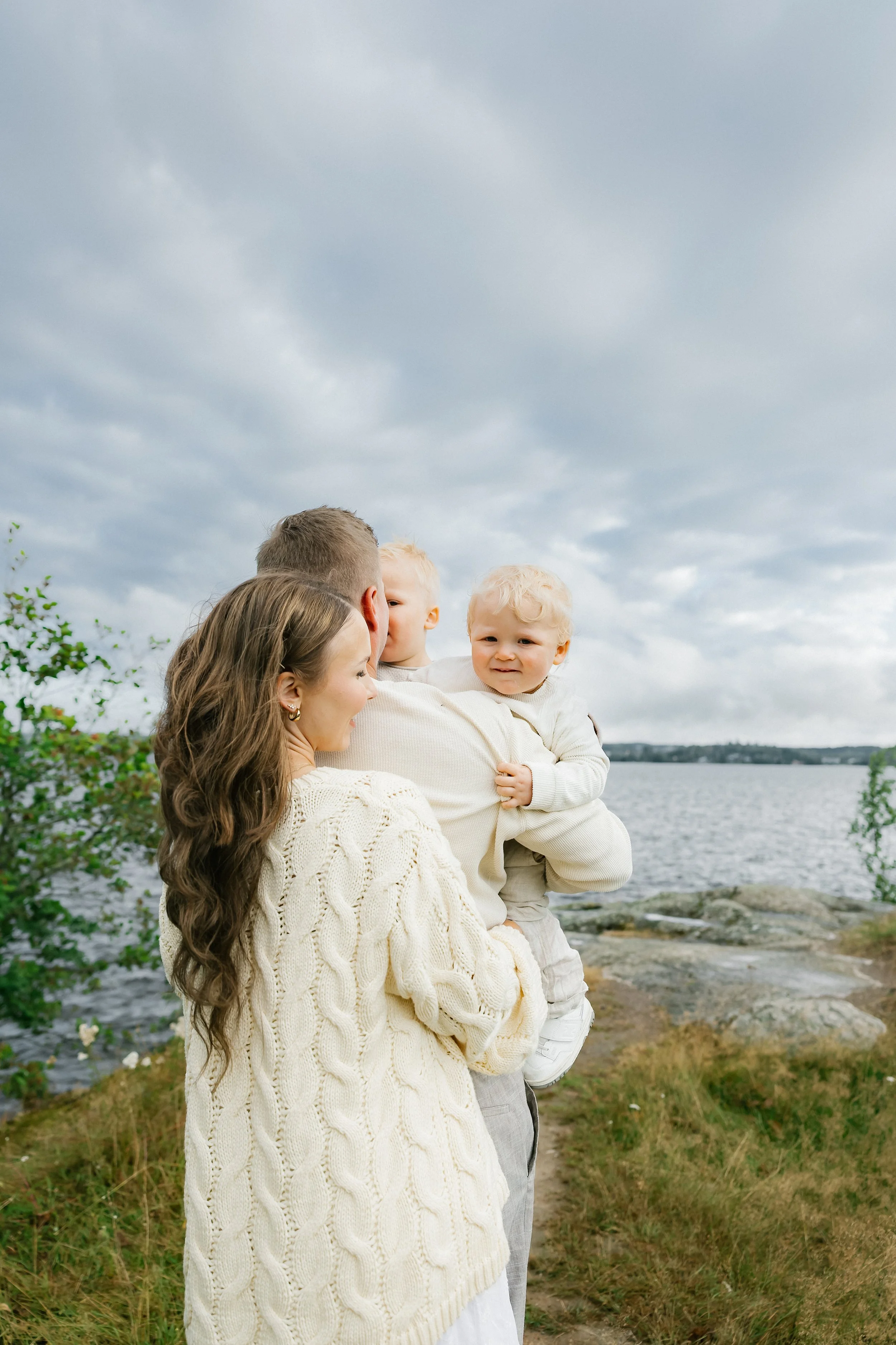 A family of four enjoying time outdoors near a body of water on a cloudy day. The mother has long, wavy brown hair and is wearing a cream-colored knitted sweater. The father is holding two young children, one with curly blonde hair and a smile, and t