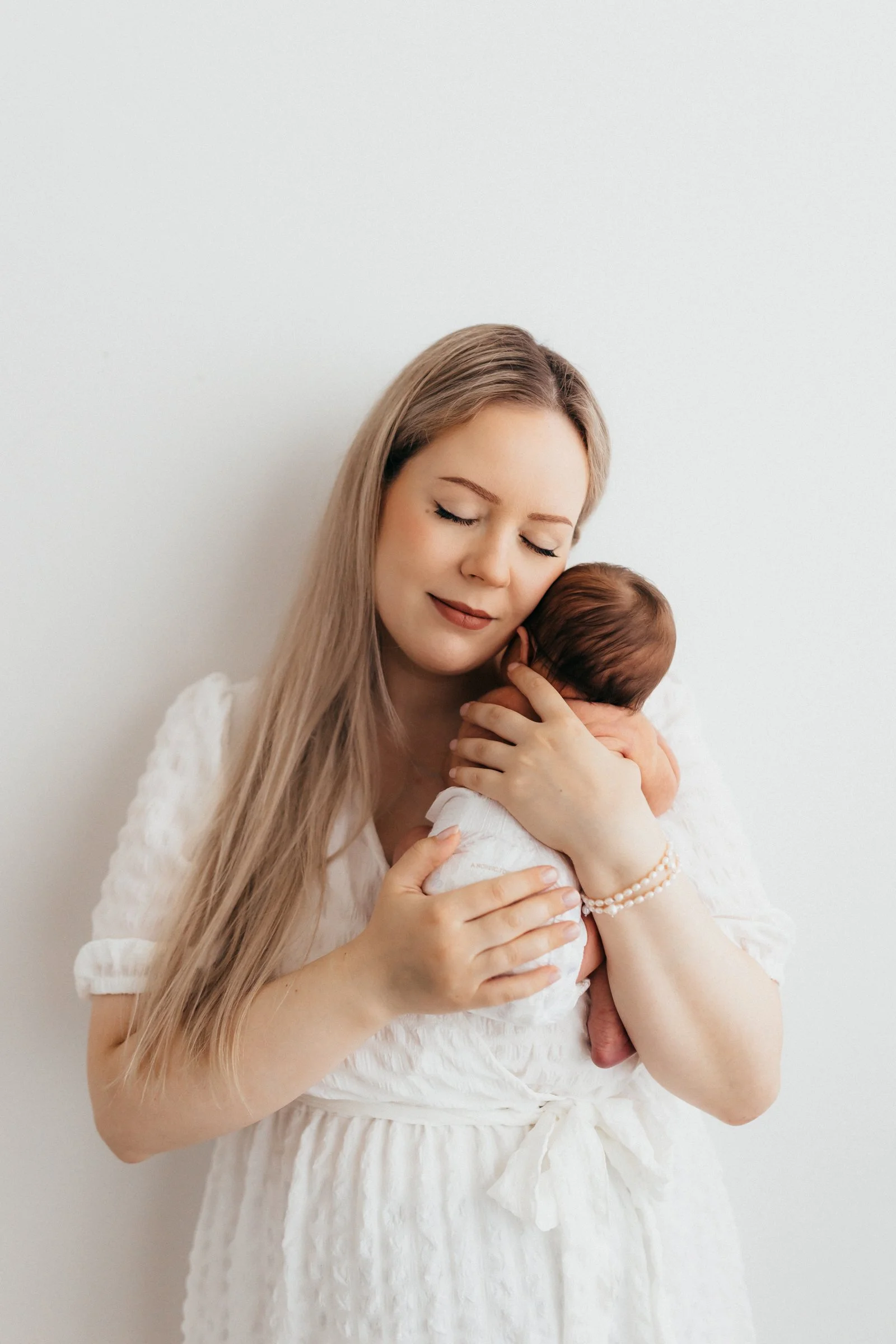 A woman with long blonde hair holding a newborn baby close to her chest, both with their eyes closed, against a plain white background.