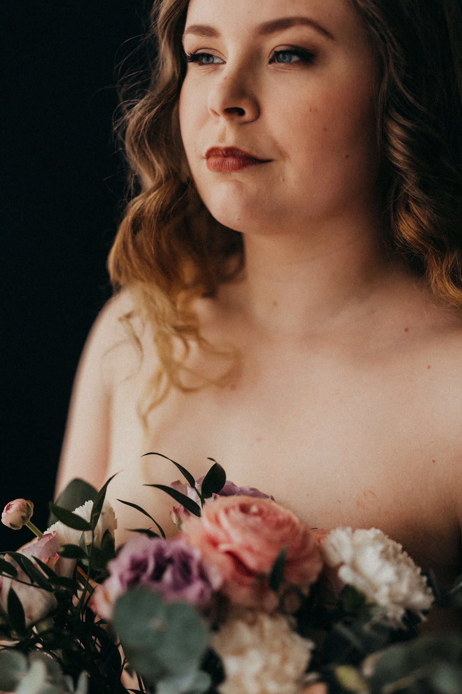 Close-up portrait of a young woman with wavy hair holding a bouquet of pink, purple, white, and peach flowers.