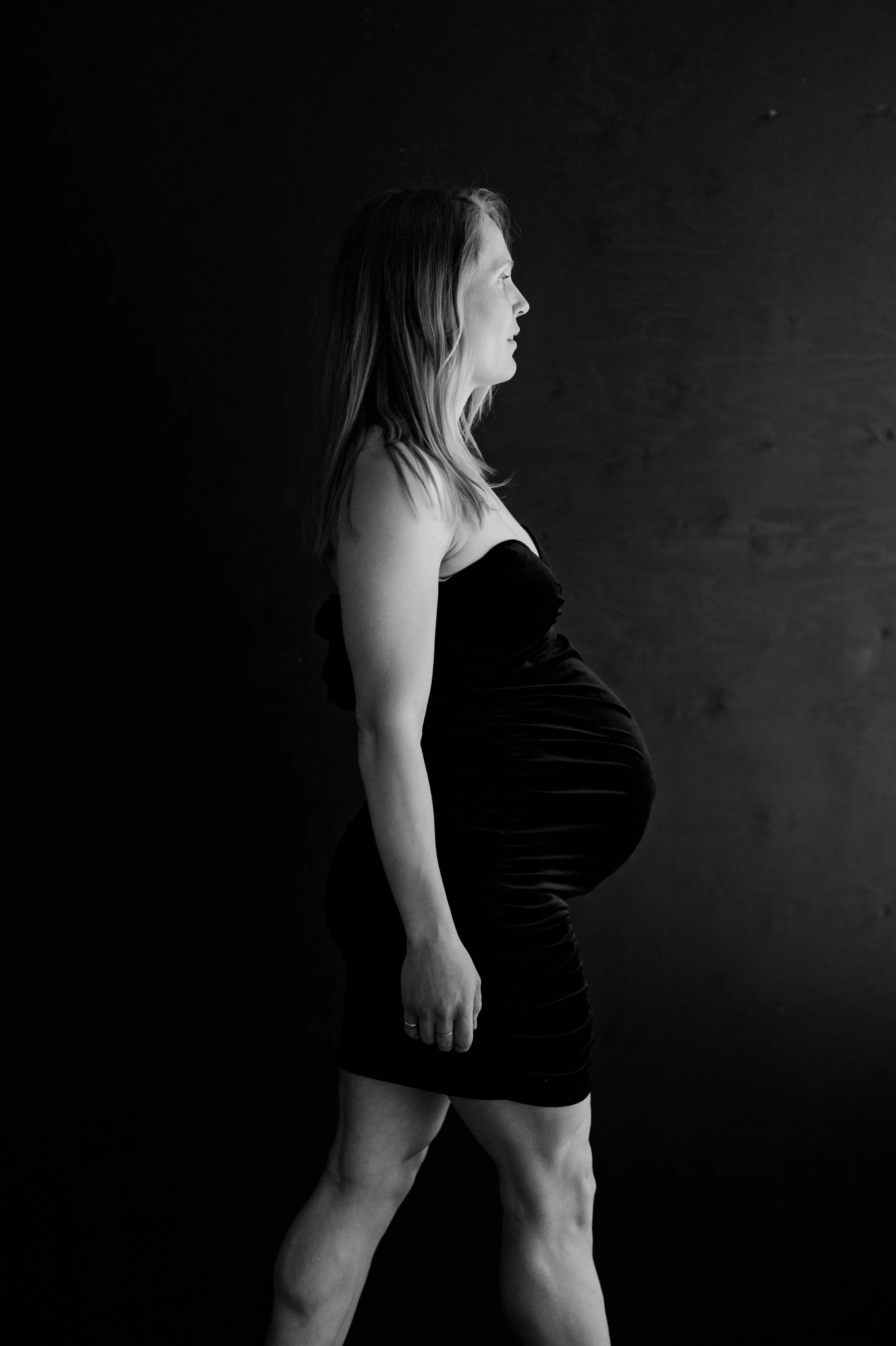 Black and white photo of a pregnant woman standing sideways against a dark background.