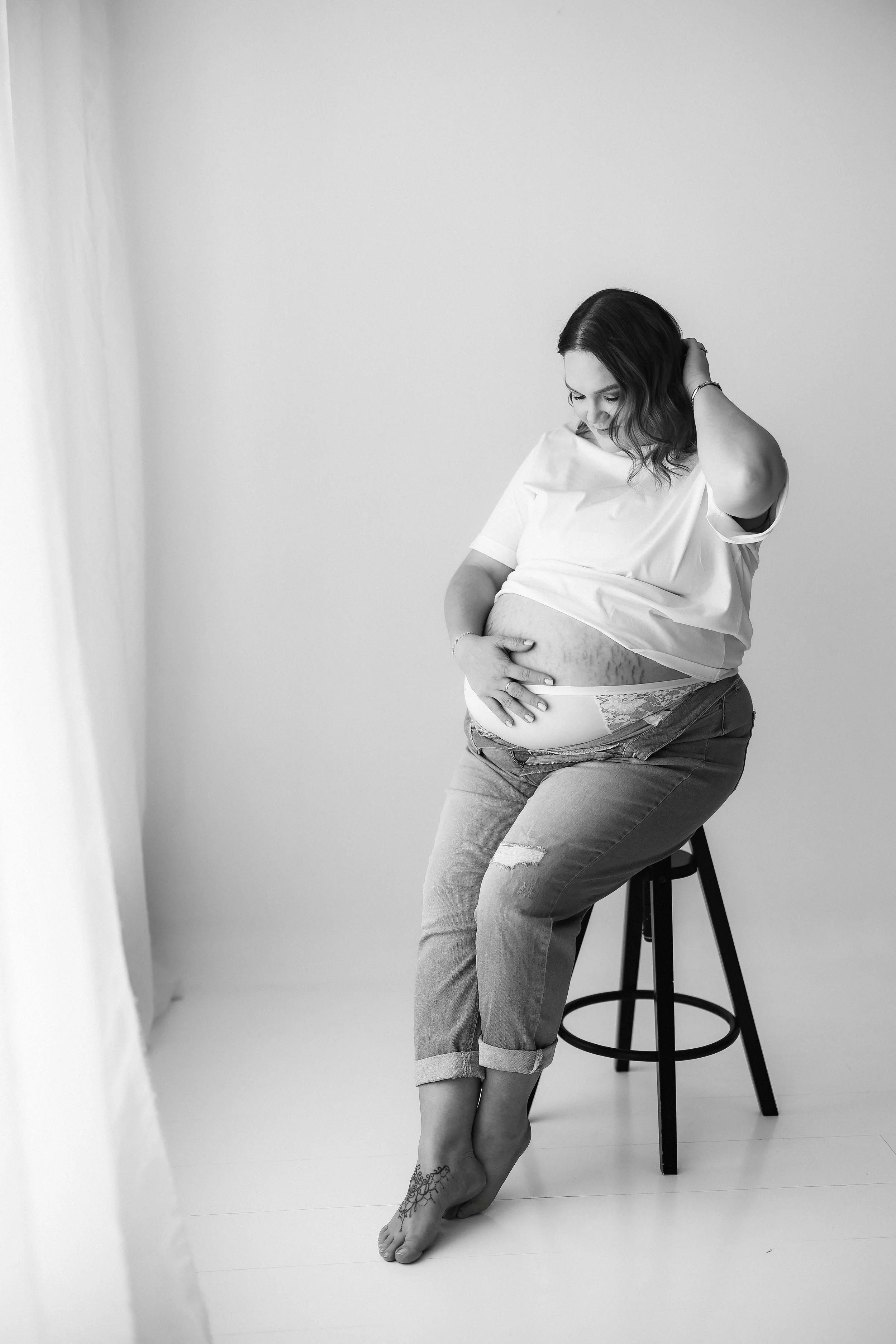 A pregnant woman sitting on a stool with her hand on her belly, looking down with a smile, in a simple white room.