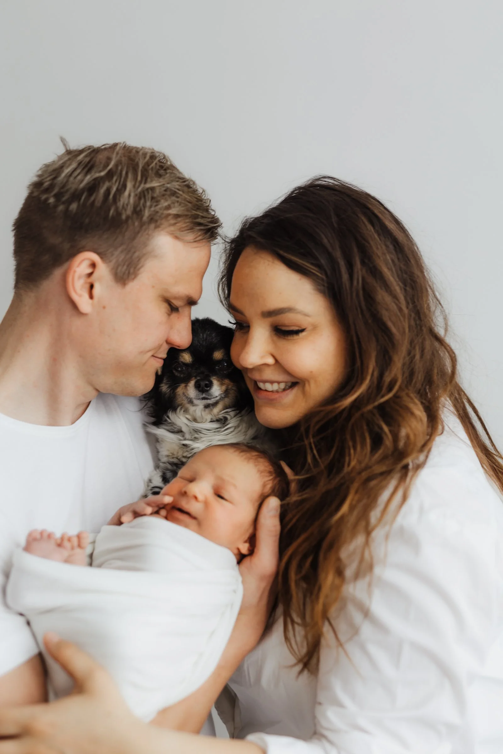A happy family of four, including a man, woman, a newborn baby, and a small dog, sharing an intimate moment together.