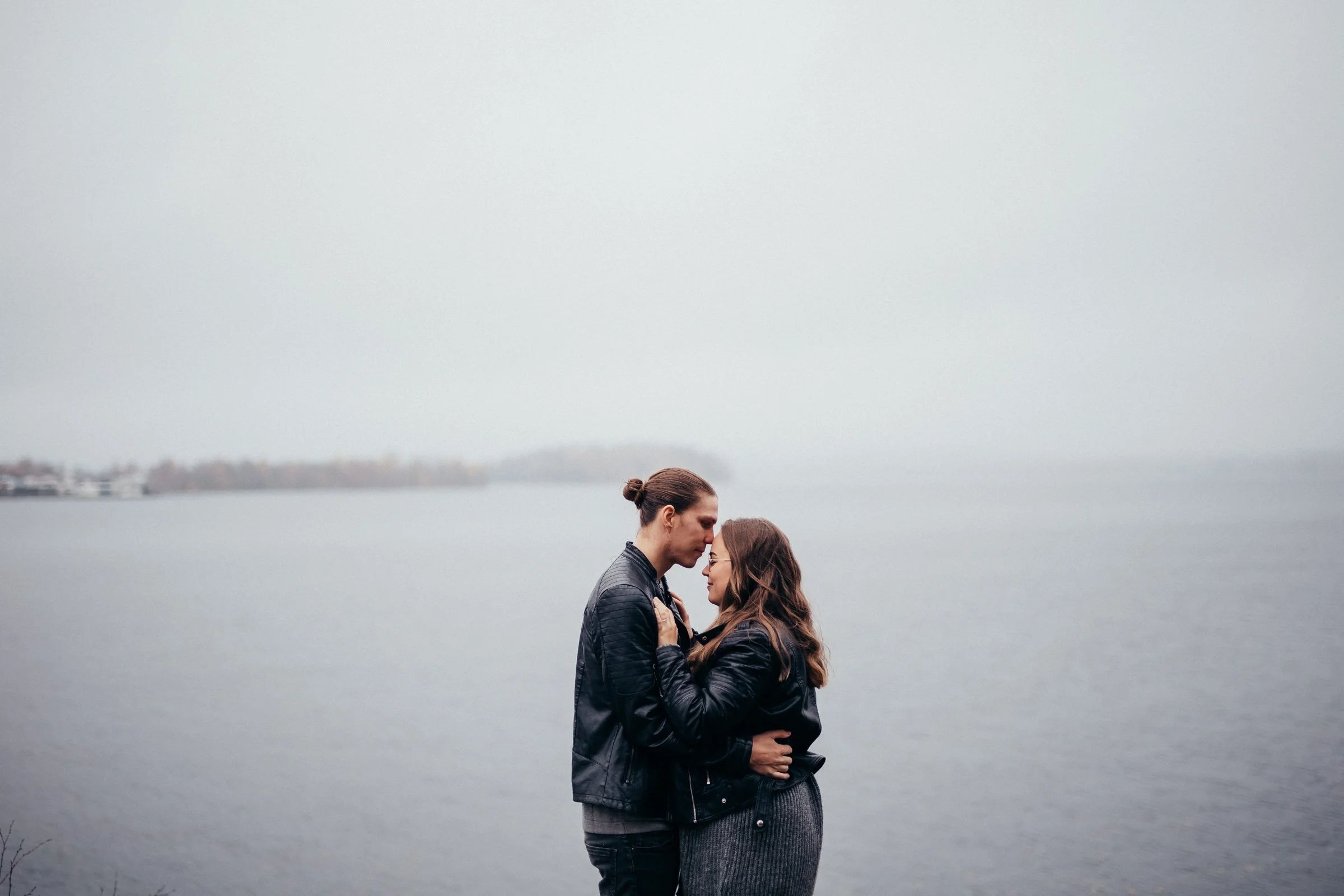 A couple standing close together by a body of water on a gray, overcast day, with their foreheads touching and holding each other.