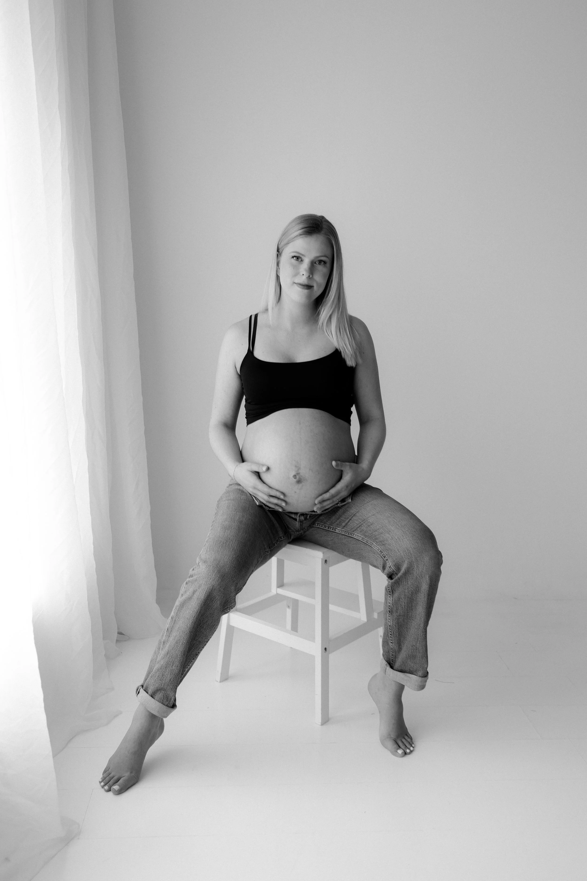 A pregnant woman with long blonde hair sitting on a white stool in a minimalistic room with a plain white wall and sheer curtains, holding her belly with both hands, wearing a black tank top and rolled-up jeans, in black and white photography.