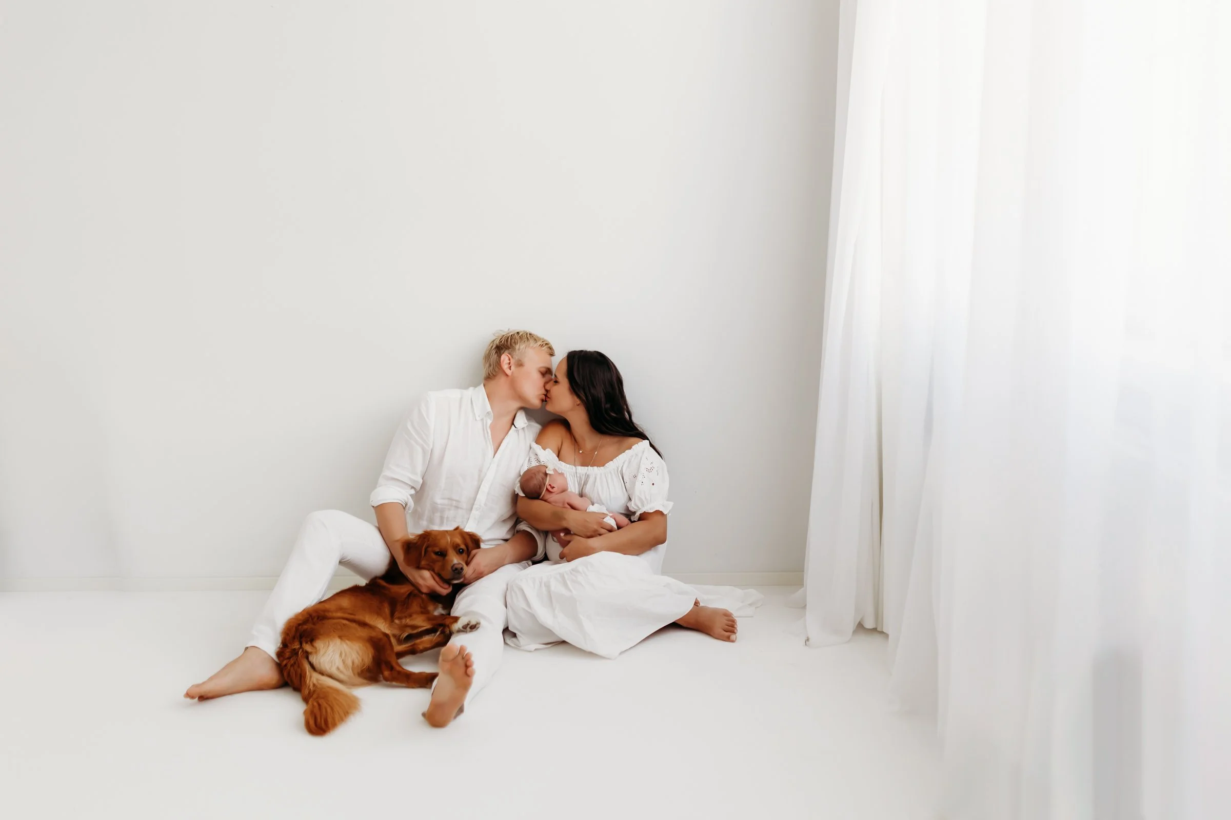 A couple, a woman with dark hair and a man with blonde hair, are sitting on a white floor against a plain white wall, kissing. The woman is holding a newborn baby, and a golden retriever is lying beside them, with its head resting on the woman's lap.
