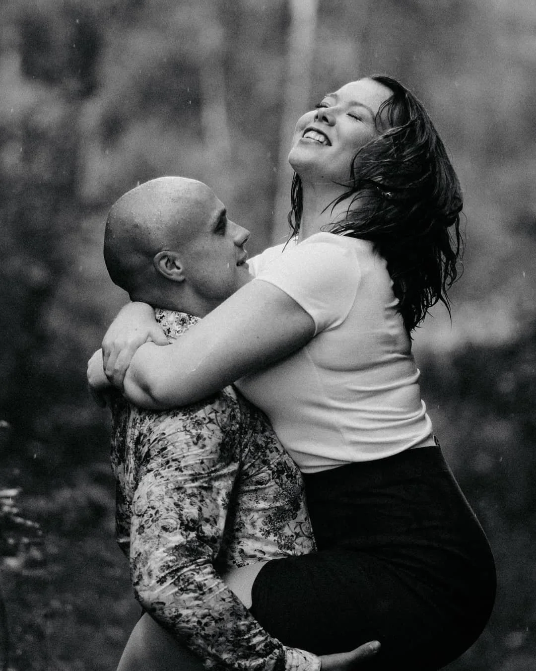 A black and white photo of a woman being lifted by a man, both smiling and laughing outdoors in the rain.