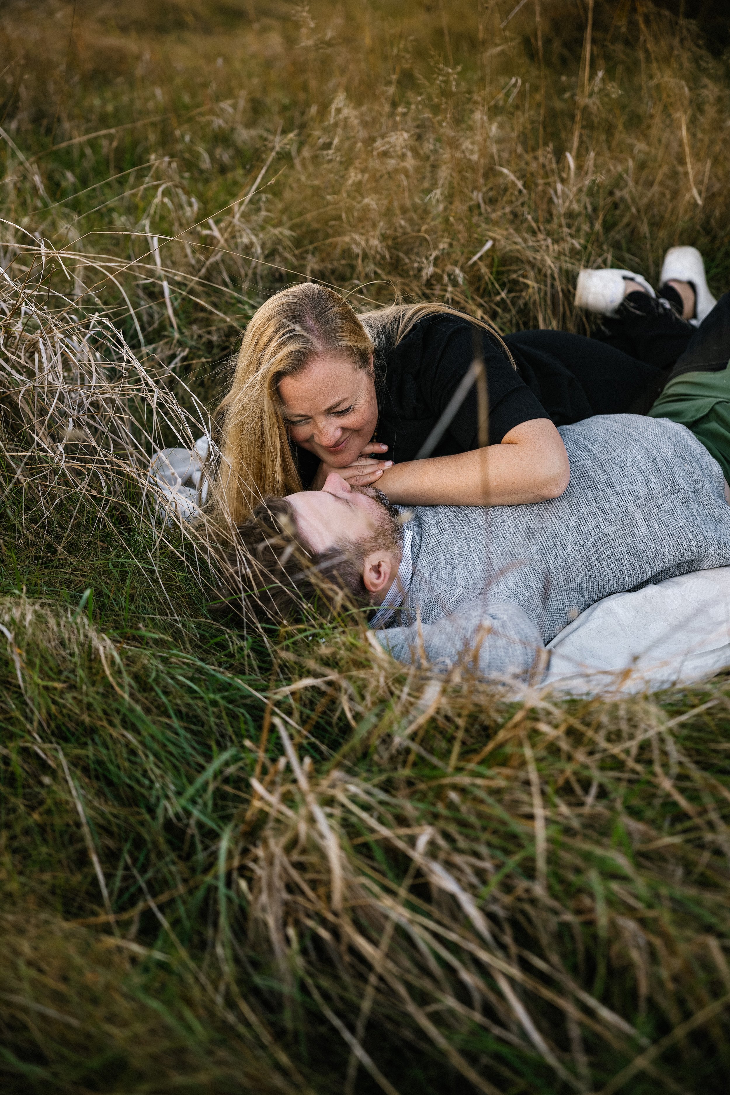 A woman and a man lying on the grass, smiling and looking at each other affectionately in an outdoor setting with grass and dried plants.
