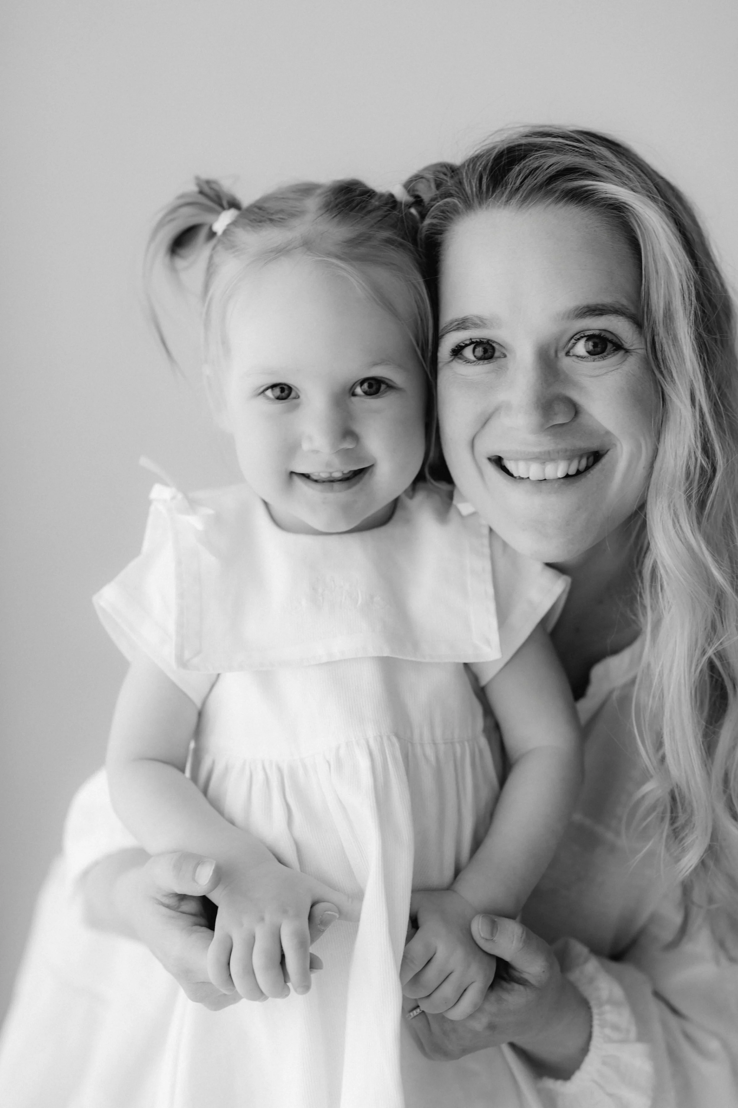 A woman and a young girl smiling and holding hands. The woman has long, wavy hair and the girl has pigtails. Both are dressed in light-colored clothing.