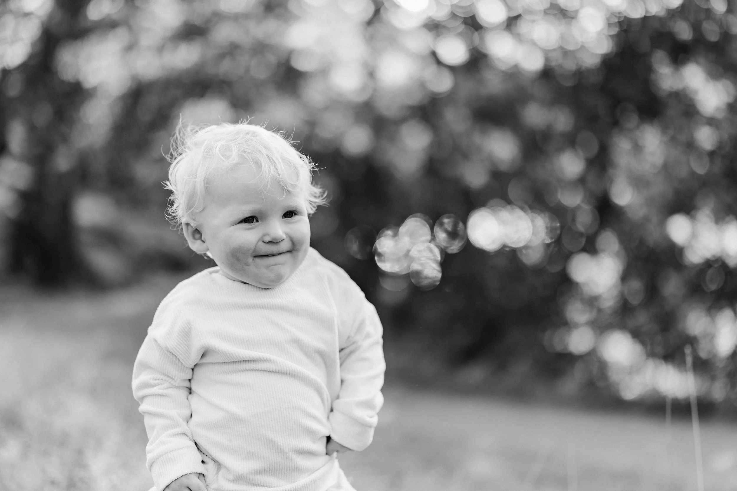 A young child with curly light-colored hair smiling outdoors in a grassy area with blurred trees in the background.