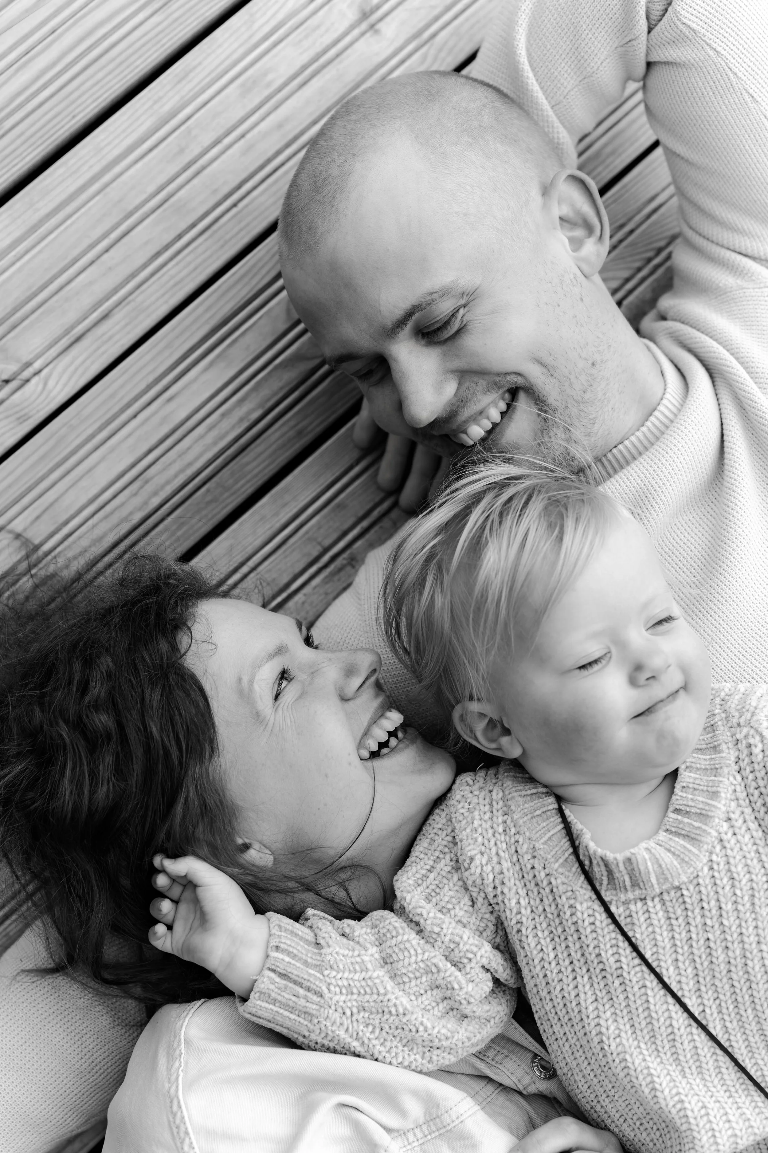 A black and white photo of a family lying on wooden planks, smiling and looking at each other.