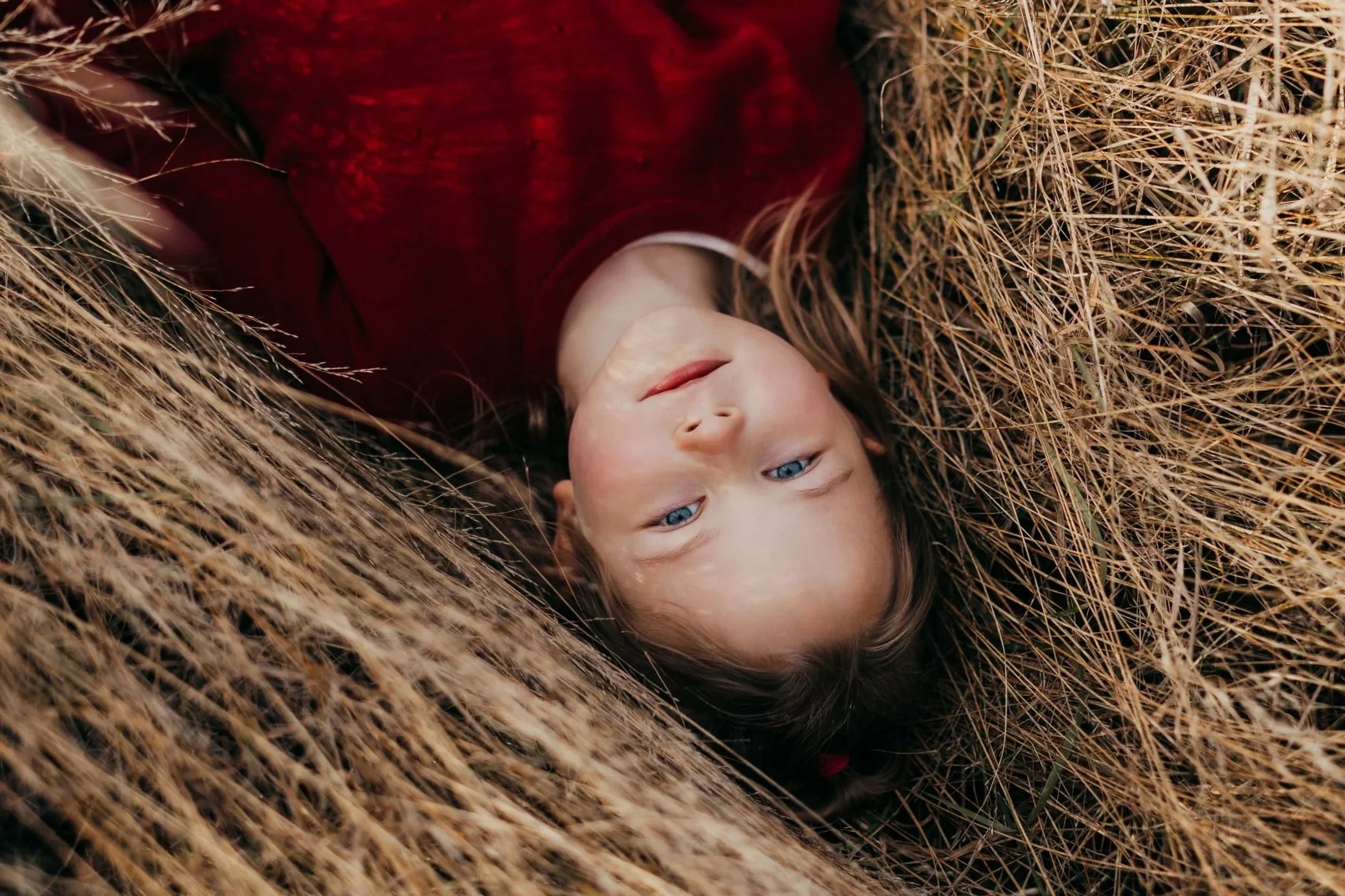 A young girl with blonde hair and blue eyes lying in a patch of dry grass, wearing a red top.