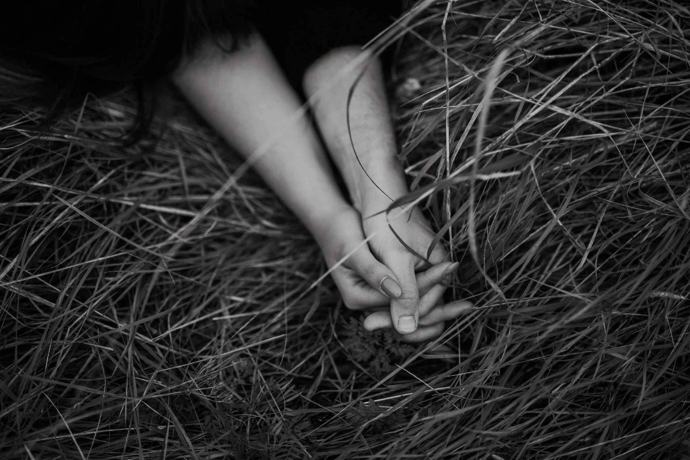 Black and white photo of a person's hands clasped together, resting on the ground amidst tall grass.