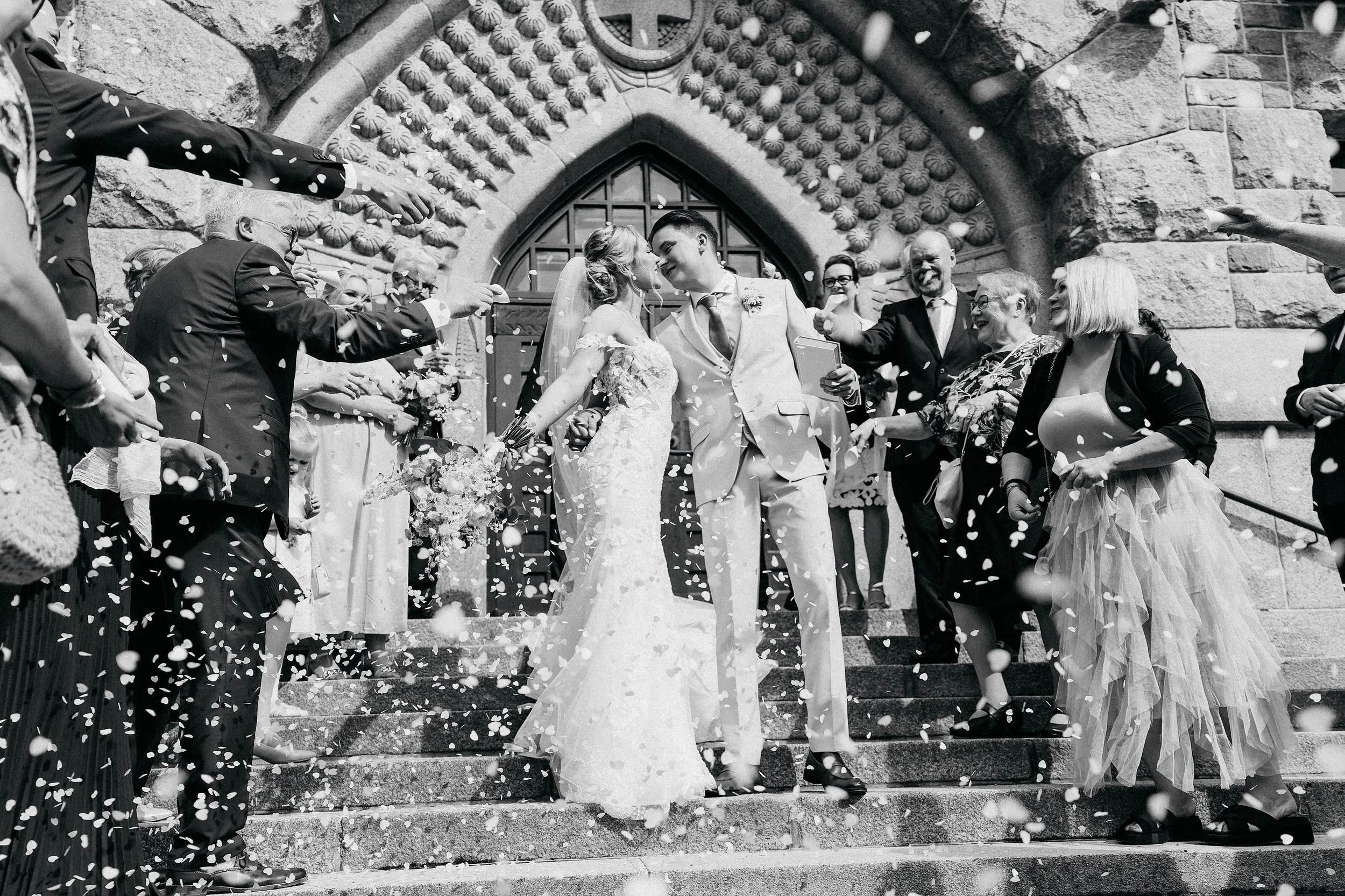 A bride and groom sharing a kiss on the steps of a church during their wedding, surrounded by guests throwing confetti.