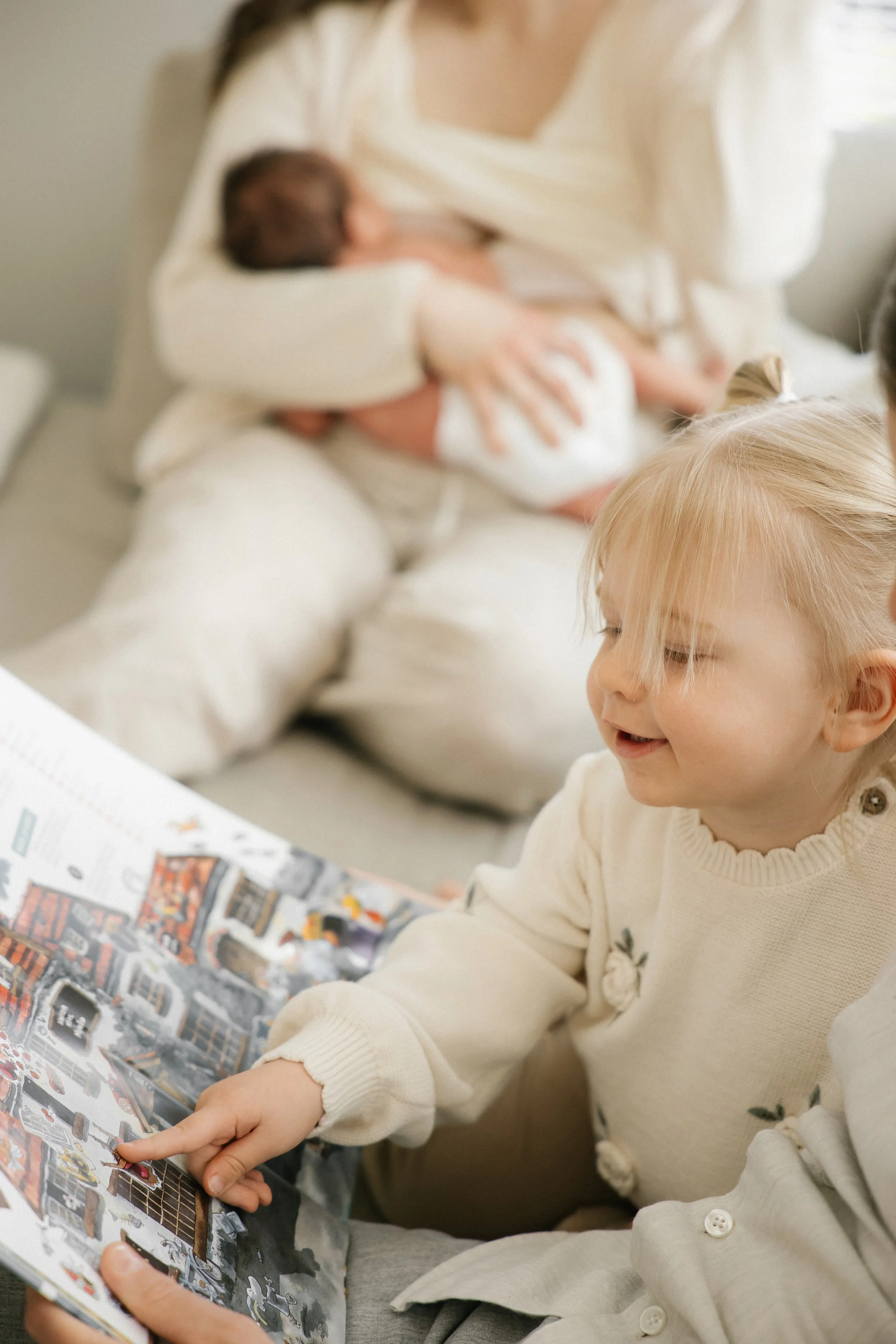 A young girl with blonde hair reading a book, with a woman holding a baby in the background.