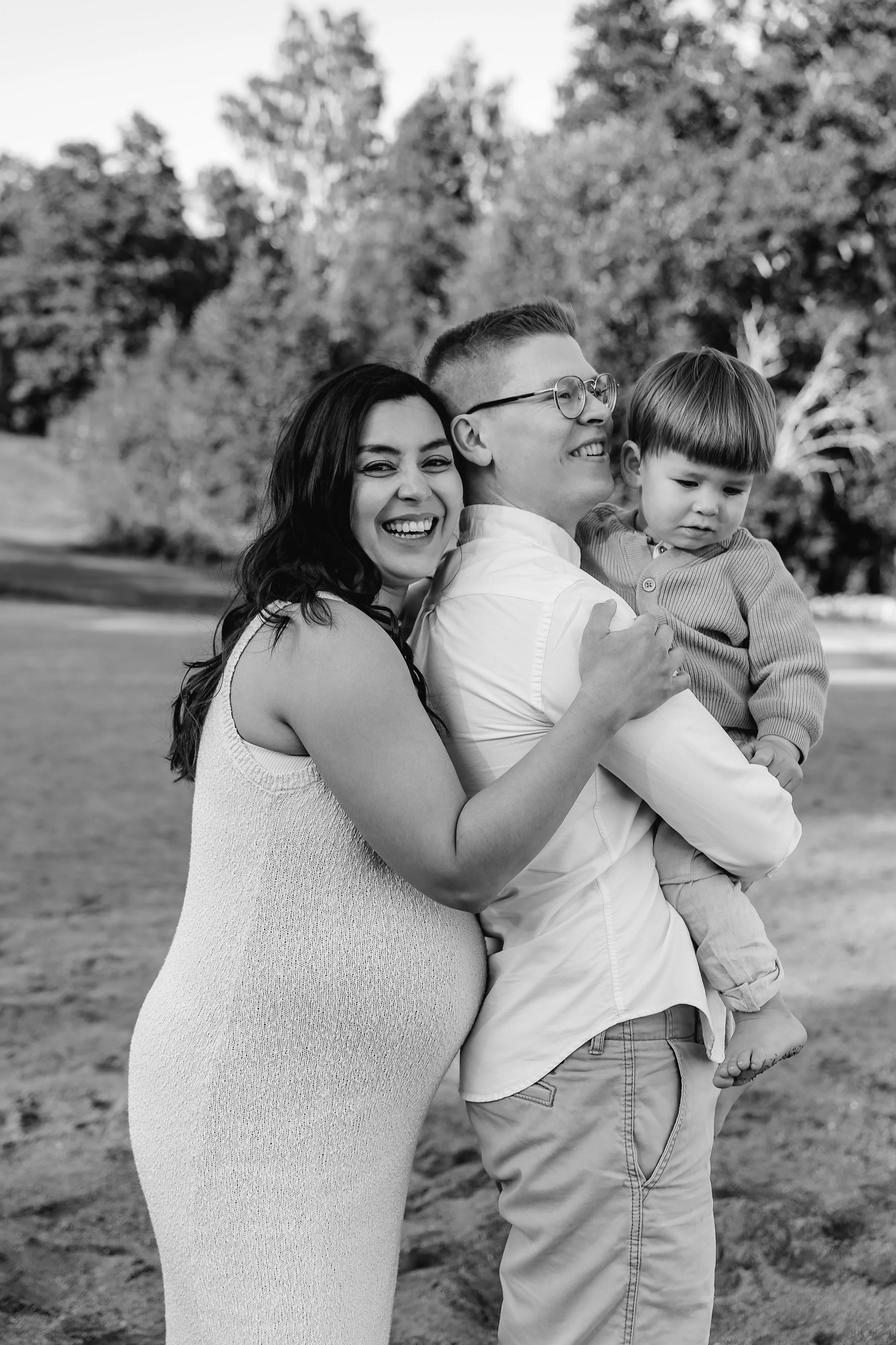 A black and white photo of a smiling woman hugging a man holding a young boy, outdoors in a park with trees in the background.