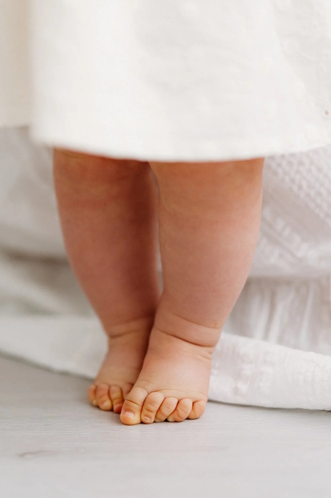 Close-up shot of baby's chubby legs and feet standing on a wooden floor, partially covered by a white fabric.