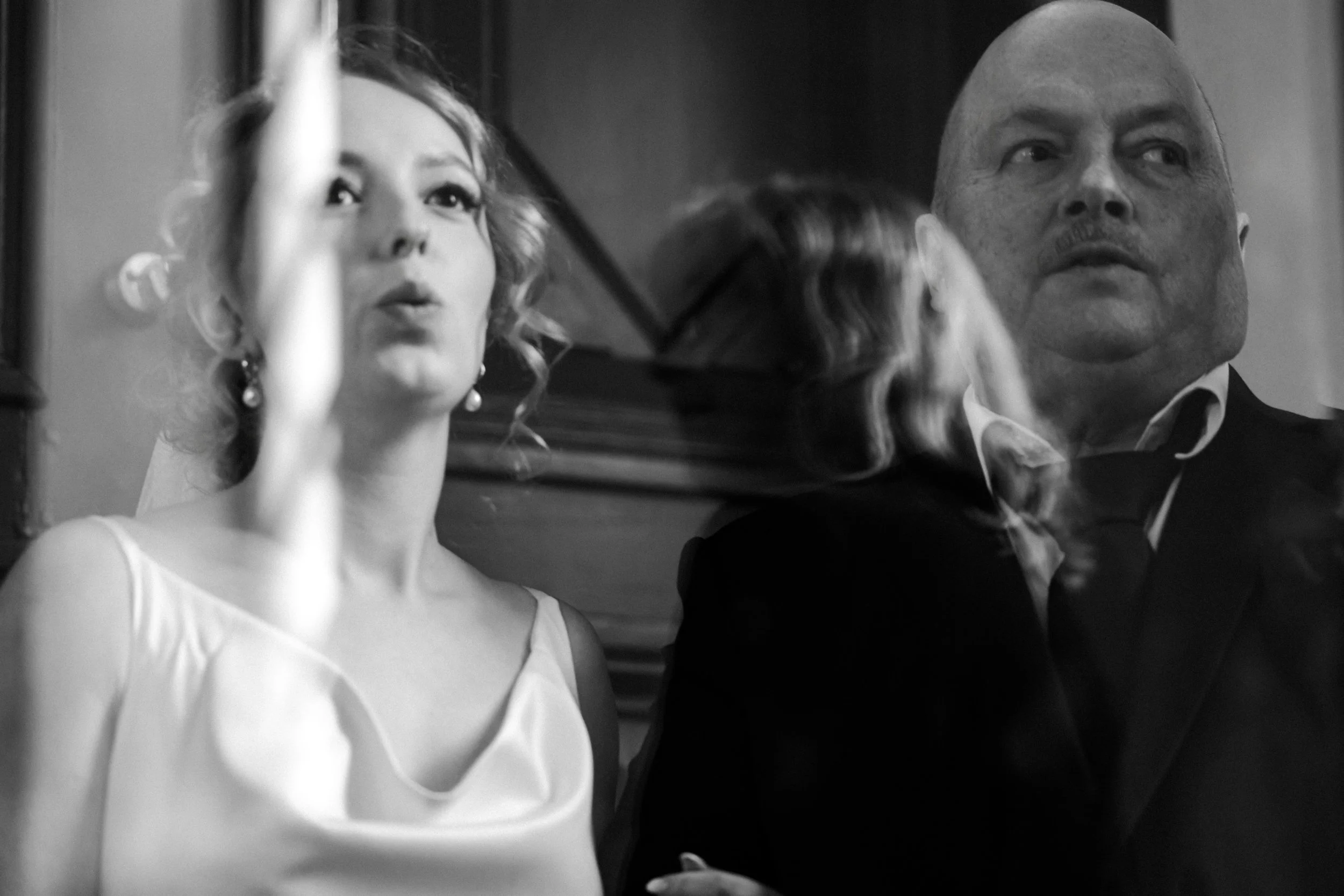 Black and white photo of a woman and a man sitting side by side, with the woman's hand on the man's shoulder. The woman has curly hair, is wearing a sleeveless dress with a wide neckline, and earrings. The man is bald, wearing a suit and tie, looking