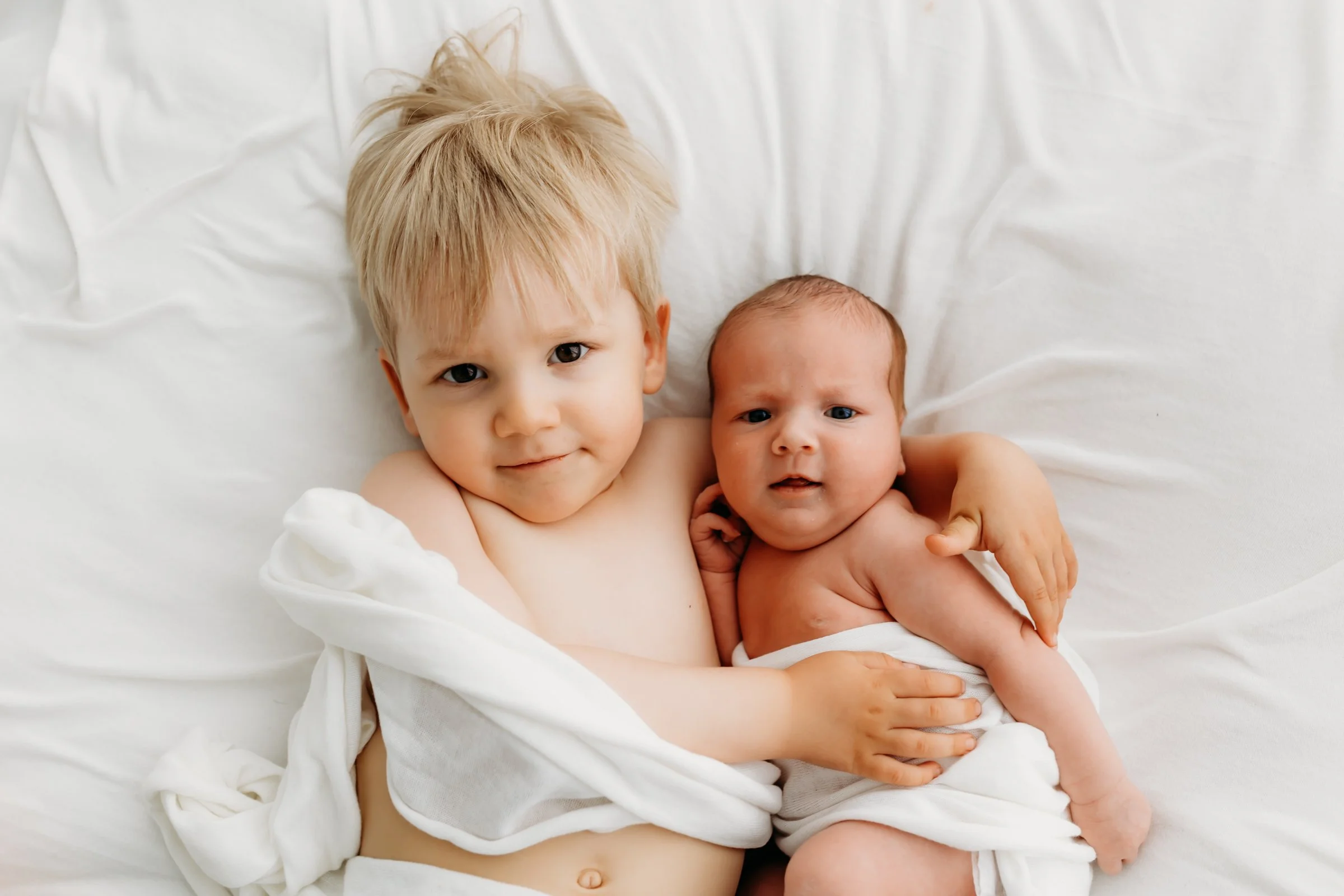 Two young children, a blonde toddler and a baby, sitting together on a white bed, wrapped partly in white sheets.