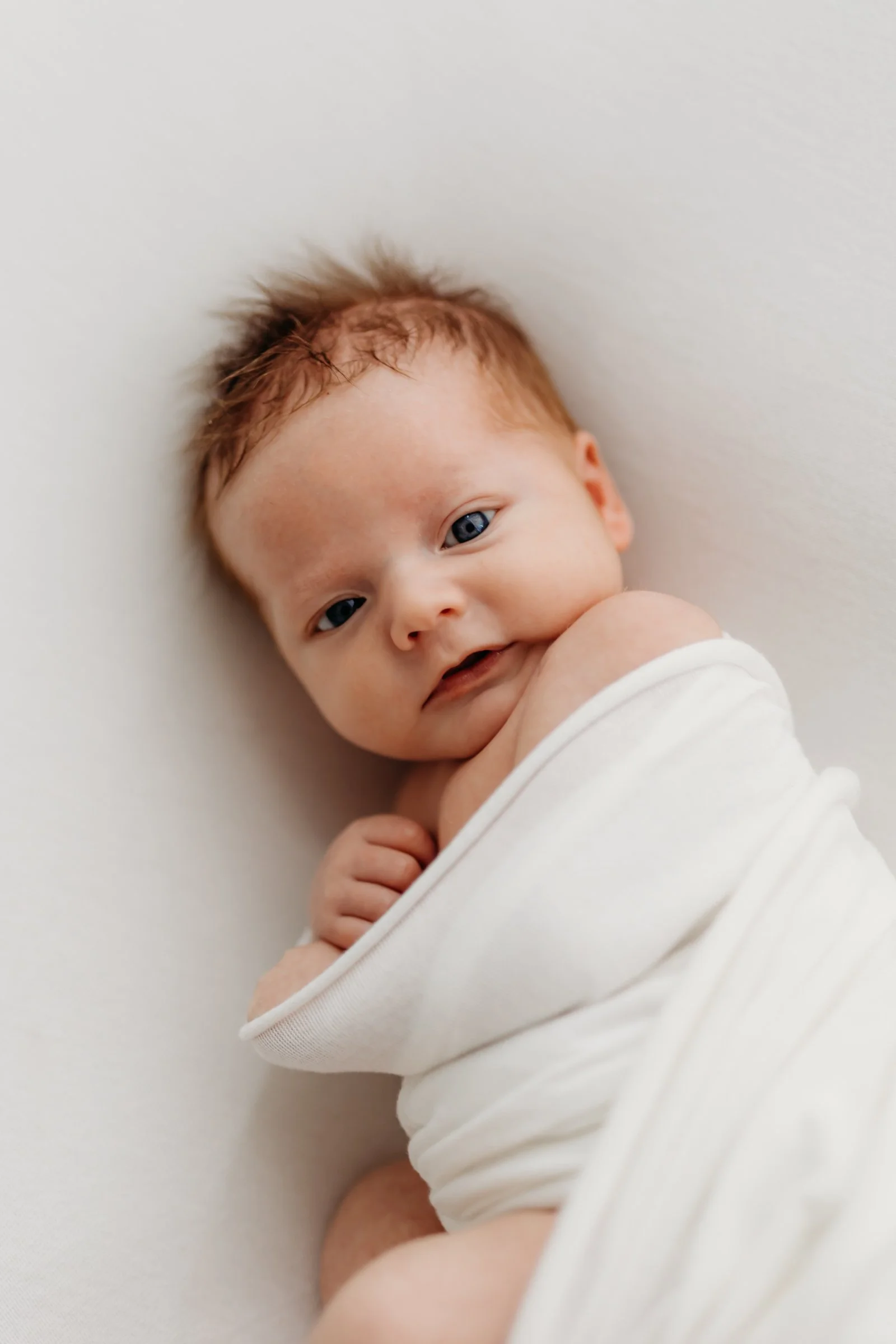 Close-up of a newborn baby with reddish hair, lying on a white surface, wrapped in a white blanket, looking at the camera.